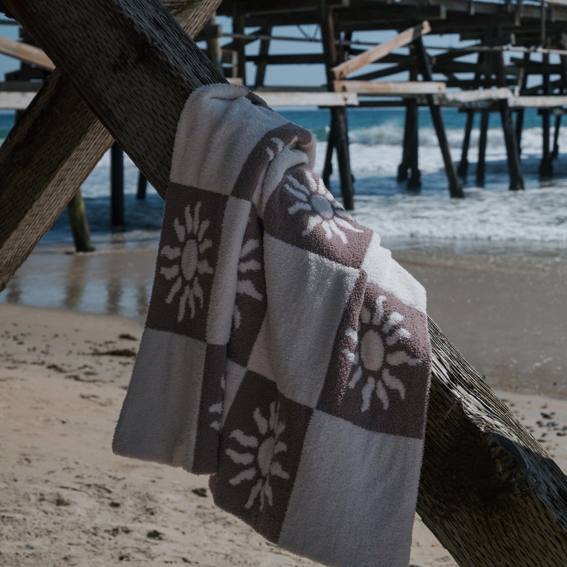 The Plush Blanket | Summer Sun by forever french baby, featuring sun patterns, is draped over a wooden pier support on a sandy beach with waves and the pier in the background under a clear sky.