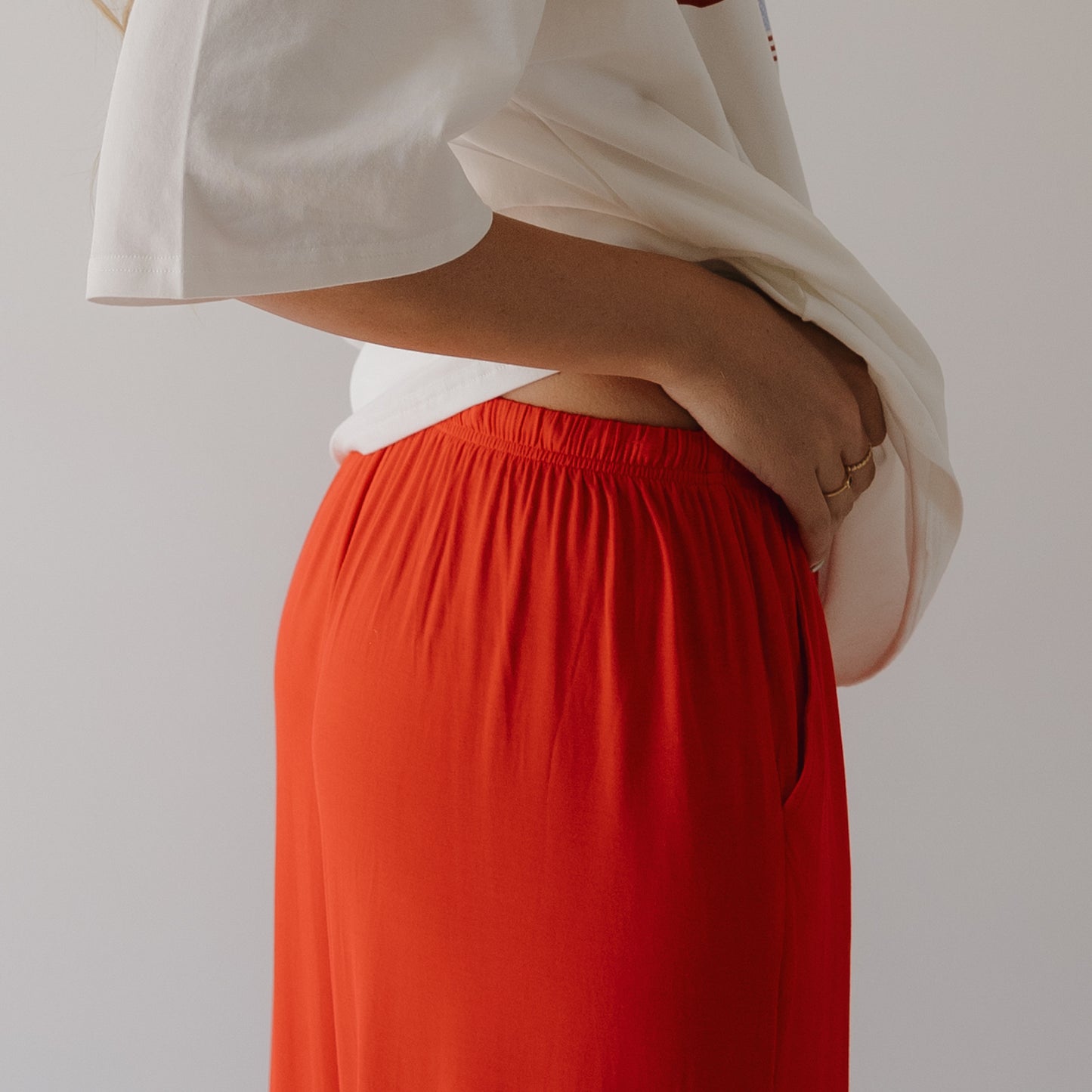 A person stands sideways in a loose white shirt and forever french baby Women's Bamboo Wide Leg Pants in Cherry Red. One hand is in the pocket, showing simple gold rings against a plain, light background.