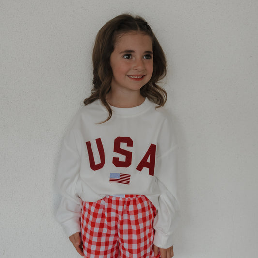 A smiling young girl with long brown hair models the forever french baby *PRE-ORDER* Child Sweatshirt | USA, featuring a red "USA" and American flag, paired with red and white checkered pants against a plain white background.