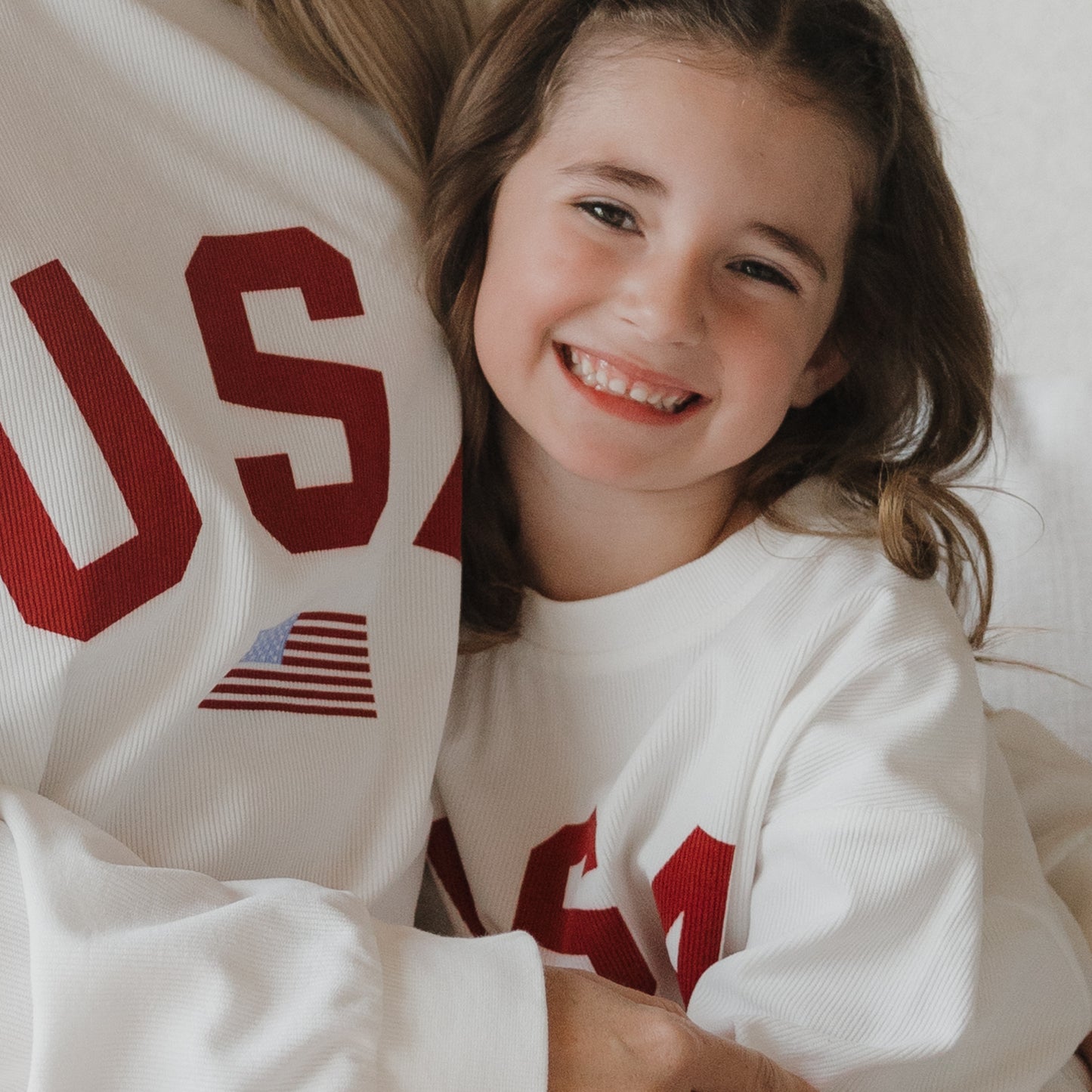 A smiling young girl in a forever french baby *PRE-ORDER* Child Sweatshirt | USA—featuring red "USA" letters and an American flag—hugs an adult, both wearing matching soft ribbed cotton family sweatshirts.