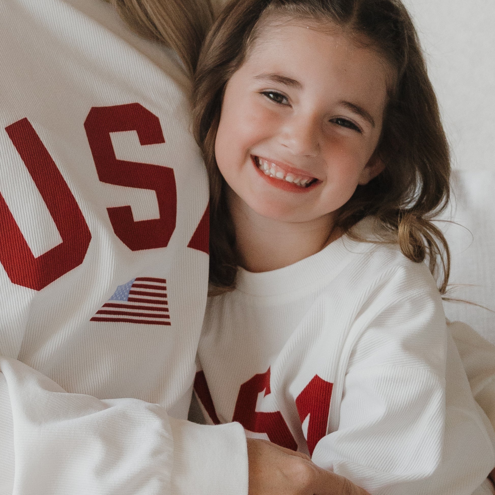 A smiling young girl in a forever french baby *PRE-ORDER* Child Sweatshirt | USA—featuring red "USA" letters and an American flag—hugs an adult, both wearing matching soft ribbed cotton family sweatshirts.