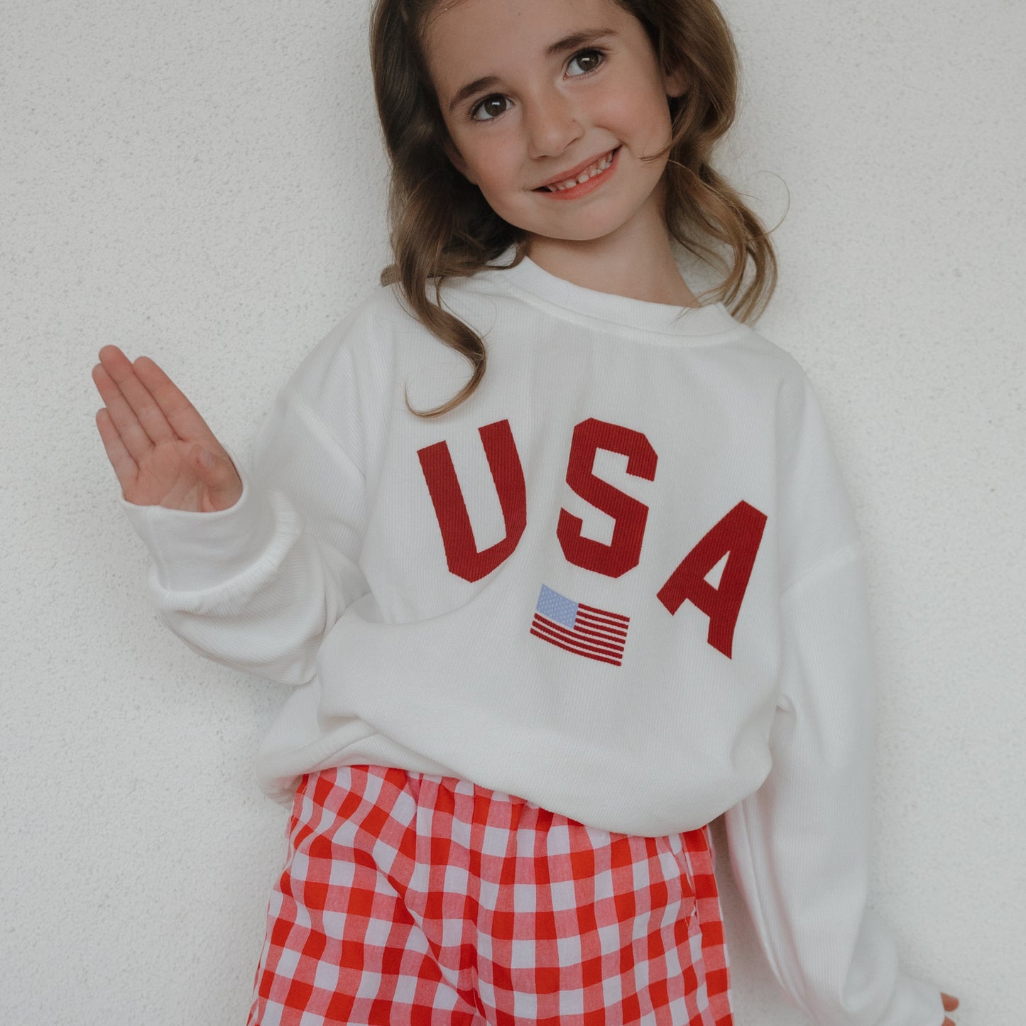 A smiling girl waves while wearing the forever french baby *PRE-ORDER* Child Sweatshirt | USA, featuring "USA" and a small flag, paired with red-and-white checkered shorts against a white wall.