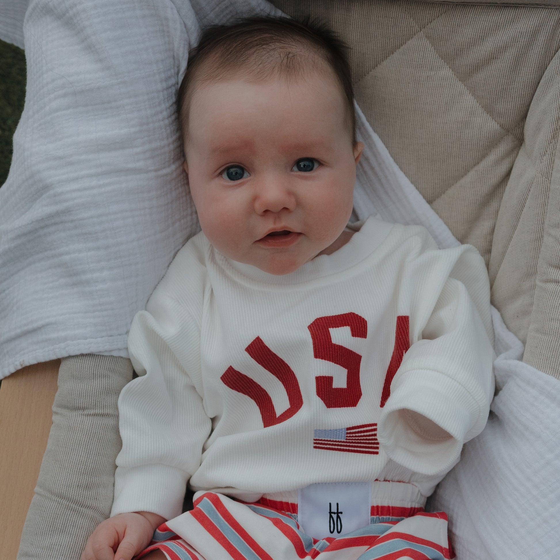 Light-skinned, blue-eyed baby in a *PRE-ORDER* Child Sweatshirt | USA by forever french baby and red-and-white striped pants, sitting in a padded seat with a white blanket.