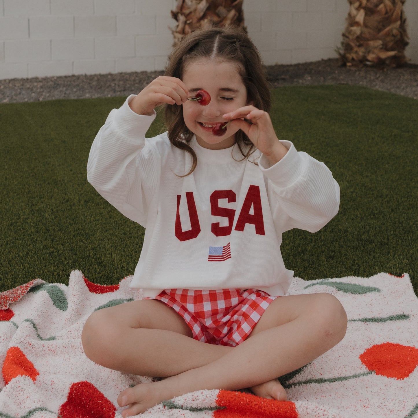 A young girl sits outside on a blanket, smiling with cherries by her eyes. She wears the forever french baby *PRE-ORDER* Child Sweatshirt | USA and red checkered shorts for a comfy, playful style.