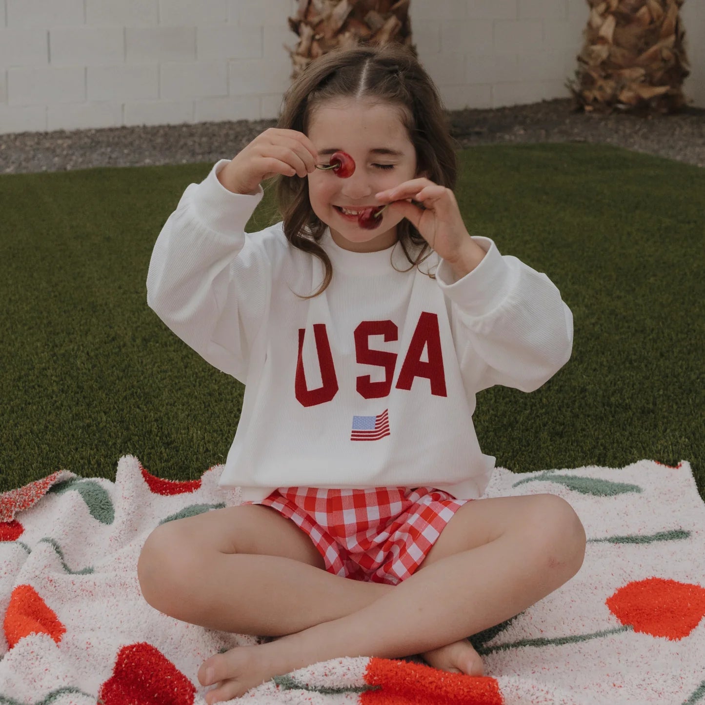 A young child sits cross-legged on a blanket outdoors, playfully holding cherries over her eyes and smiling, dressed in a white ribbed cotton forever french baby Child Sweatshirt | USA (MIS-PRINT) *FINAL SALE* and red checkered shorts.