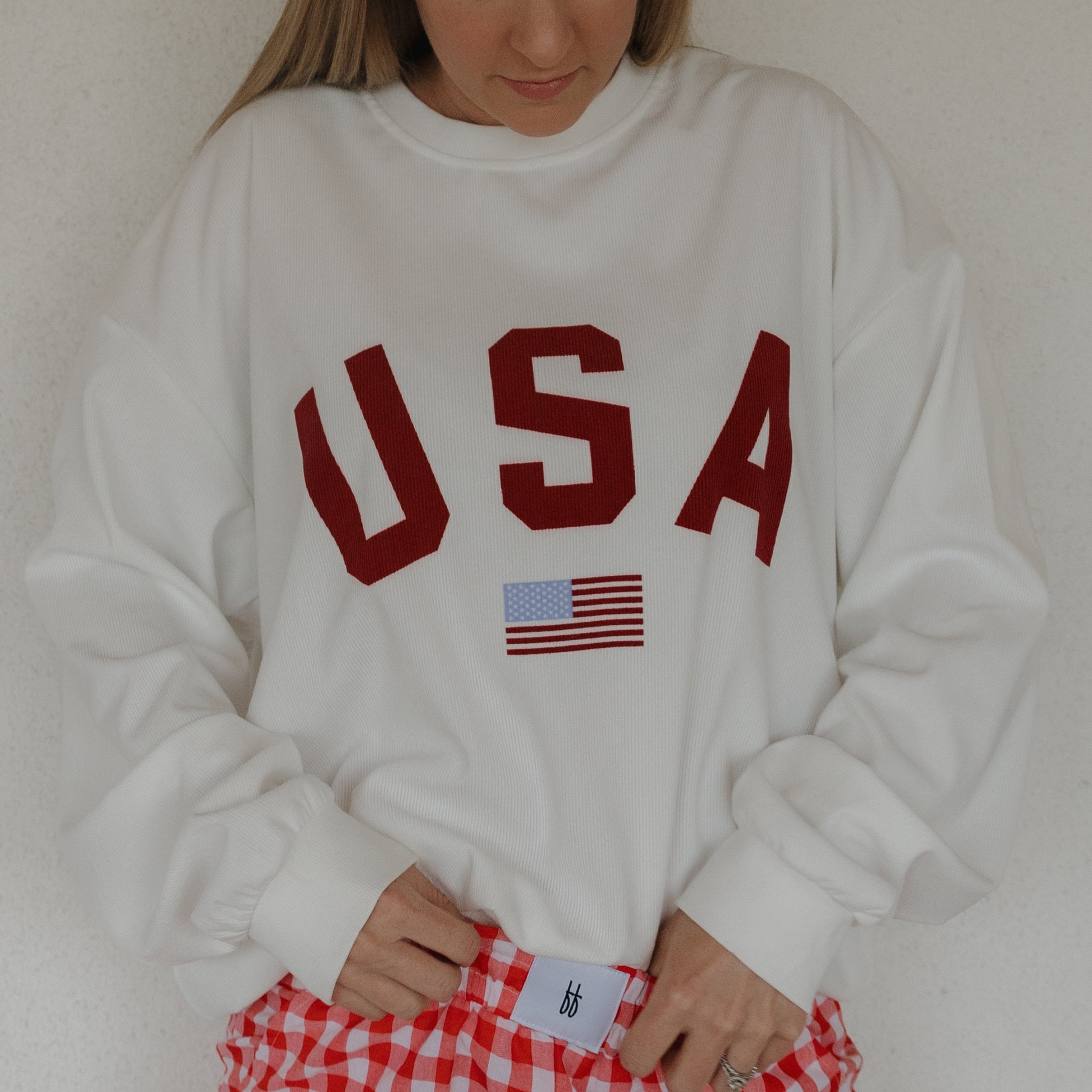 A person wears the forever french baby *PRE-ORDER* Adult Sweatshirt | USA, a relaxed fit white top with bold red "USA" and an American flag, paired with red and white checkered pants. Their face is partly out of frame; matching sweatshirts may be available.