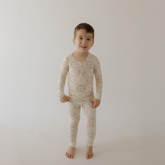 A young boy with short brown hair and a slightly uncertain expression stands barefoot on a light floor against a white wall, wearing forever french baby’s Bamboo Two Piece Pajamas | Cottage Quilt with a geometric pattern.