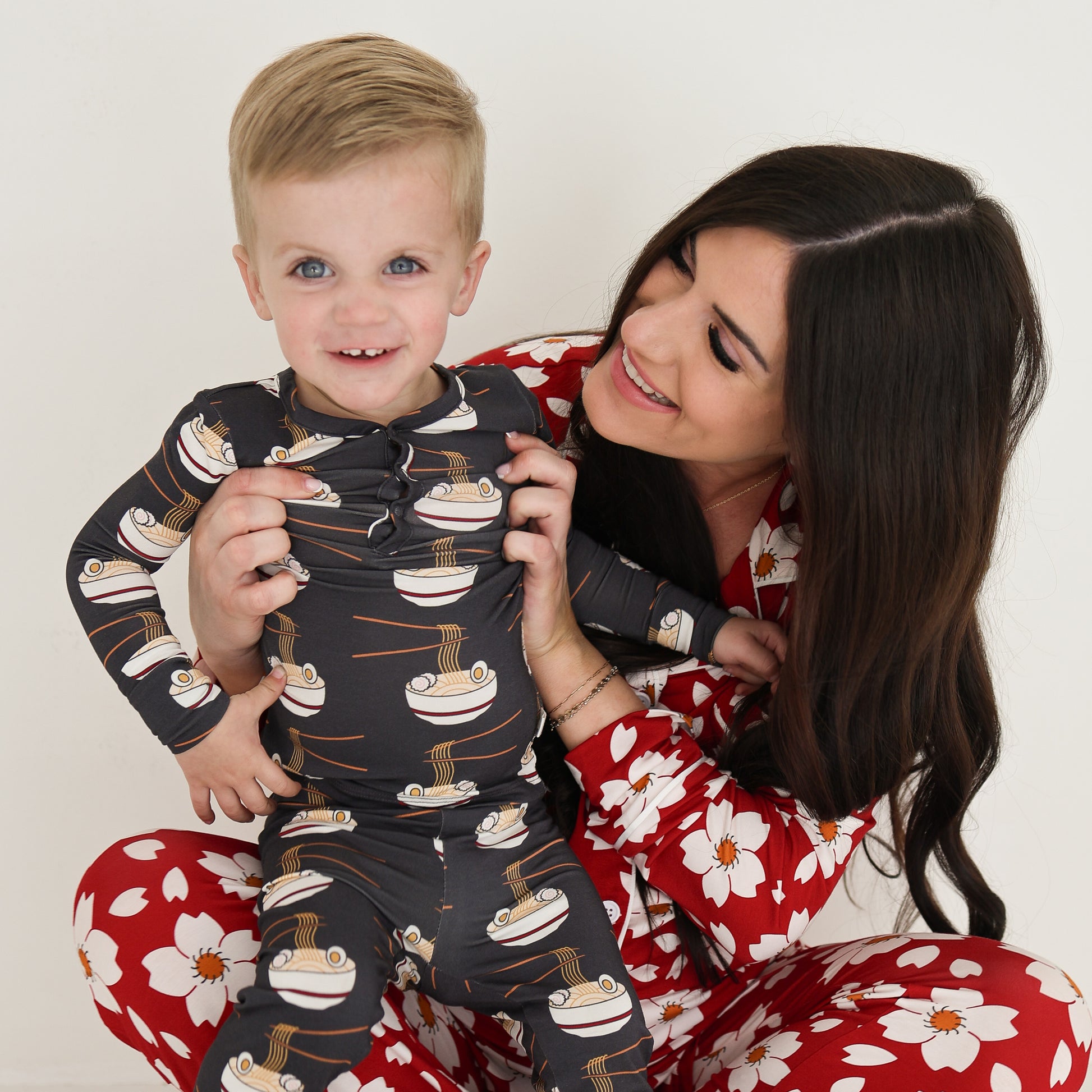 A smiling woman in red floral bamboo pajamas hugs a happy boy in forever french baby Bamboo Two Piece Pajama | Ramen. Sitting against a white background, they enjoy the cozy, hypo-allergenic comfort of these kids pajamas.
