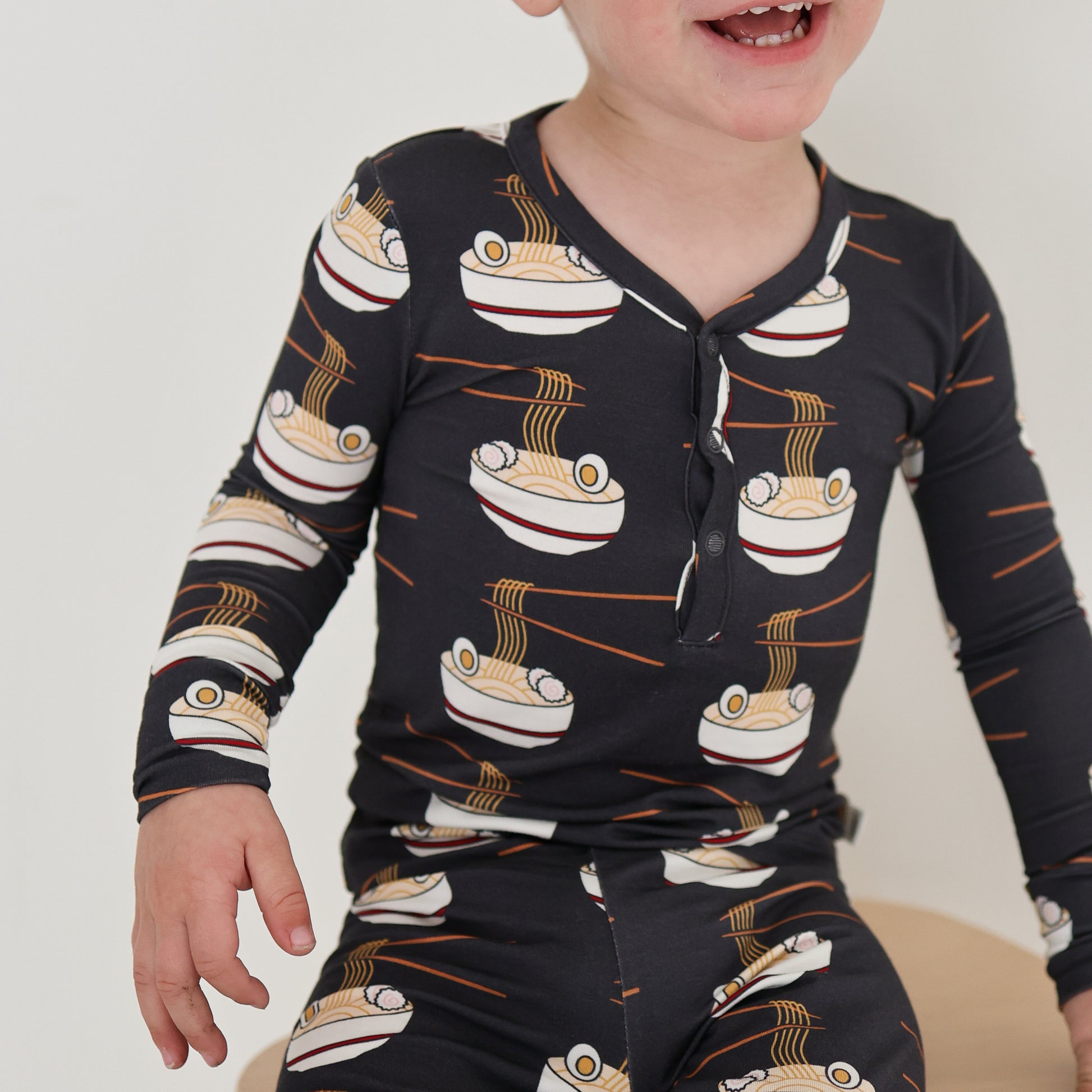 A young child in forever french baby's Bamboo Two Piece Pajama | Ramen, featuring a playful noodle bowl and chopsticks print, sits smiling with their mouth open. The focus is on the child's torso and lower face.