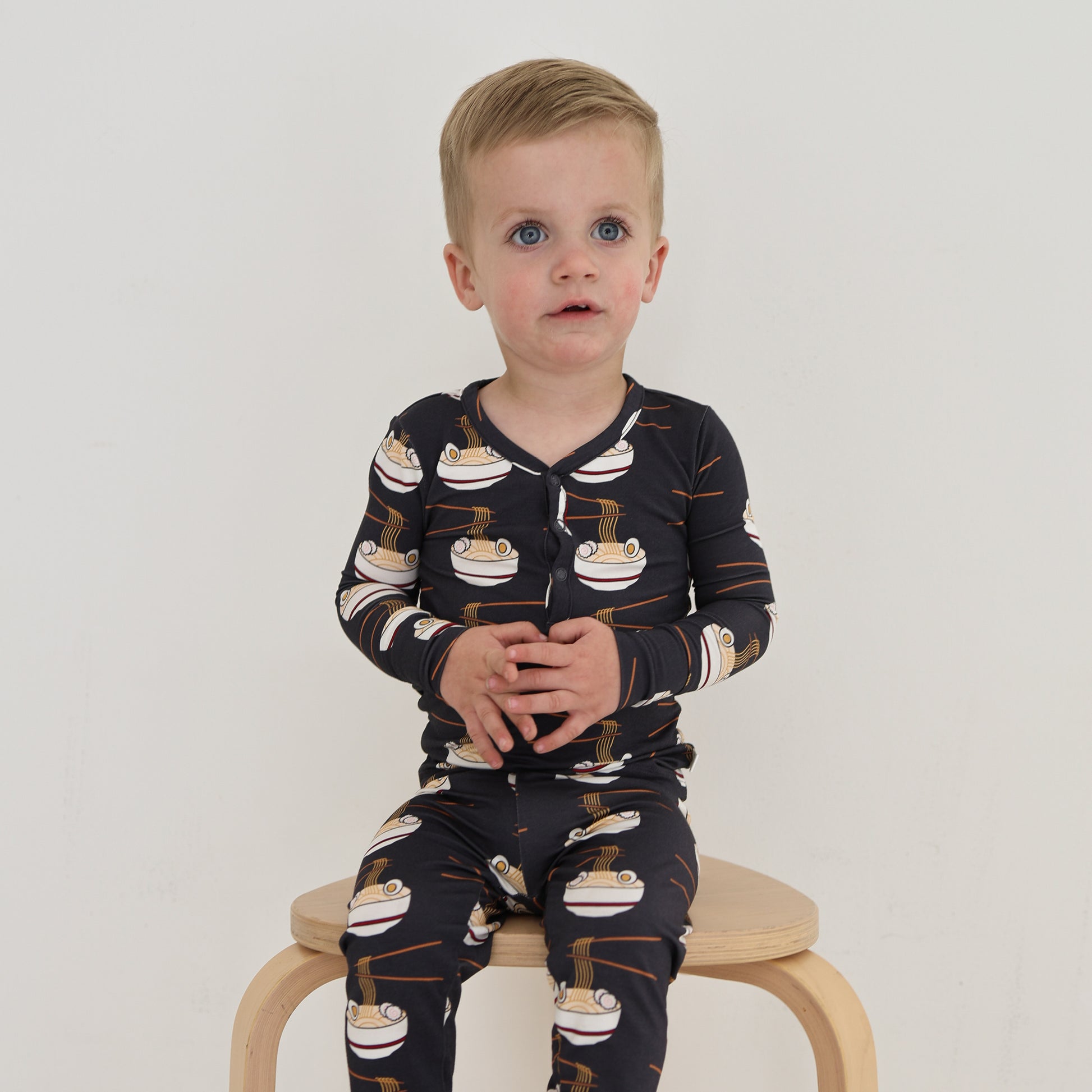 A young child with short blonde hair wears the forever french baby Bamboo Two Piece Pajama | Ramen, featuring a ramen bowl pattern, while seated on a wooden stool against a plain white background.