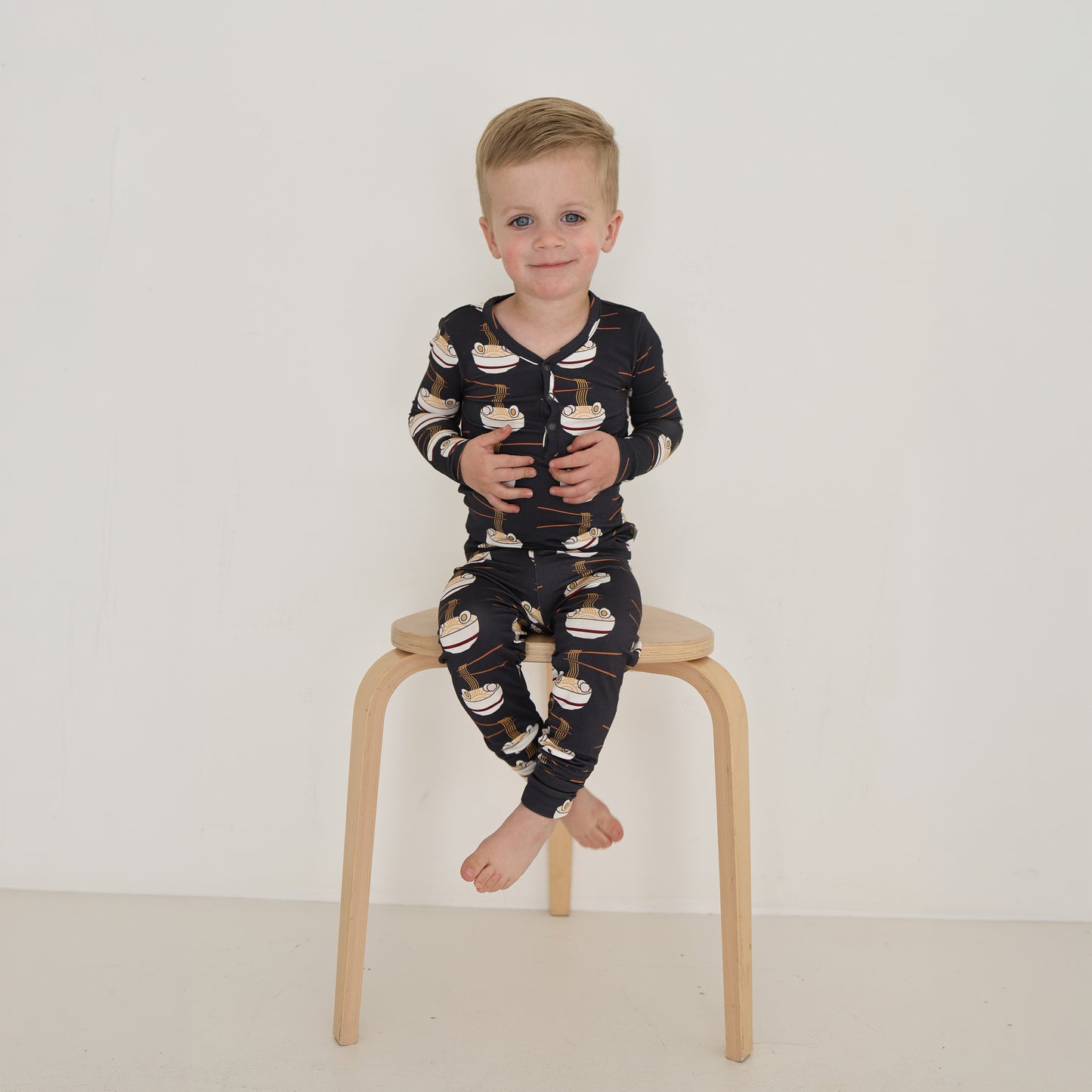A young child with short blond hair sits barefoot on a wooden stool, wearing forever french baby's Bamboo Two Piece Pajama in the Ramen print, against a plain white background.