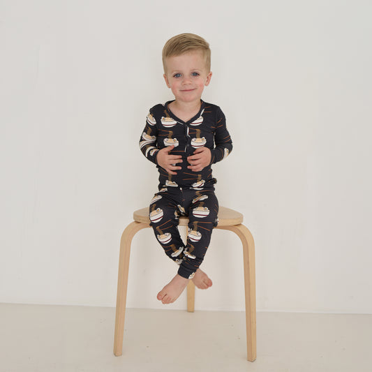 A young child with short blond hair sits barefoot on a wooden stool, wearing forever french baby's Bamboo Two Piece Pajama in the Ramen print, against a plain white background.