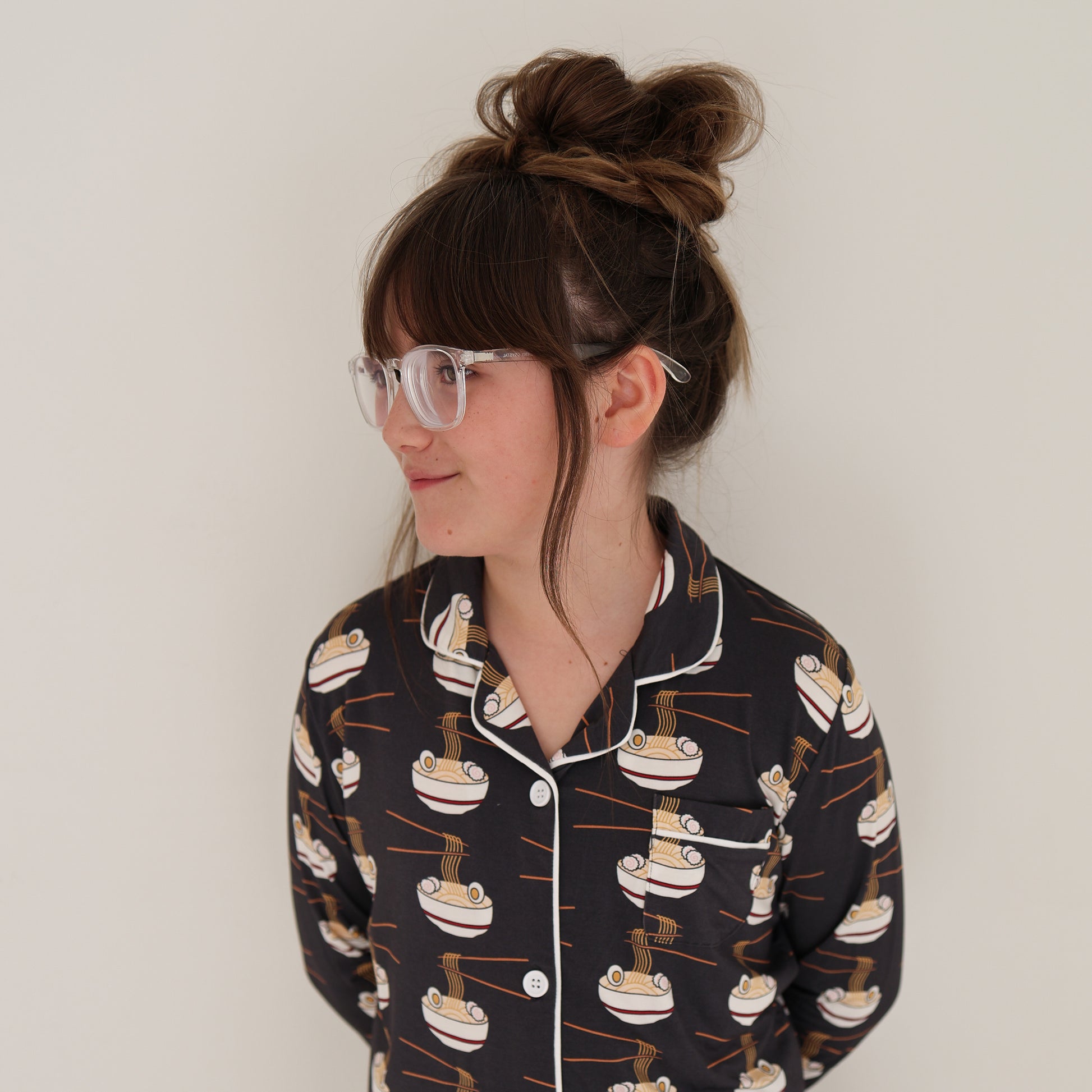 A young girl with brown hair in a messy bun and clear glasses smiles to the side, wearing the forever french baby Girl's Bamboo Button Up Lounge Set | Ramen, against a plain white background.