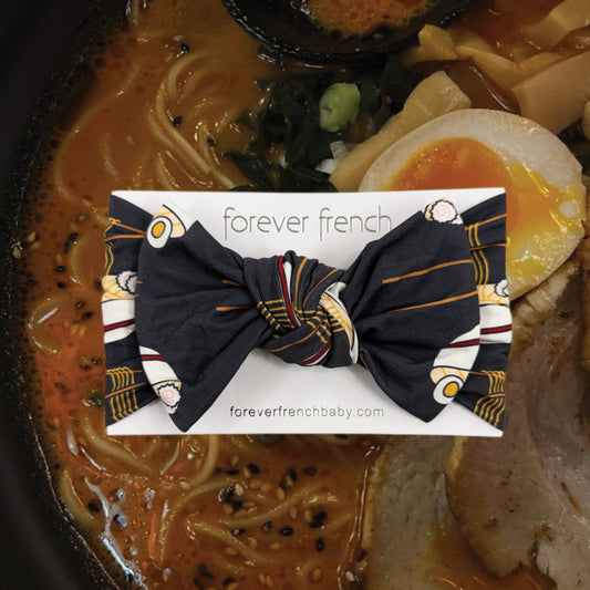 The Bamboo Head Wrap | Ramen from forever french baby, featuring ramen noodle and egg prints, is displayed attached to a branded card above a bowl of ramen with broth, noodles, egg, and pork.