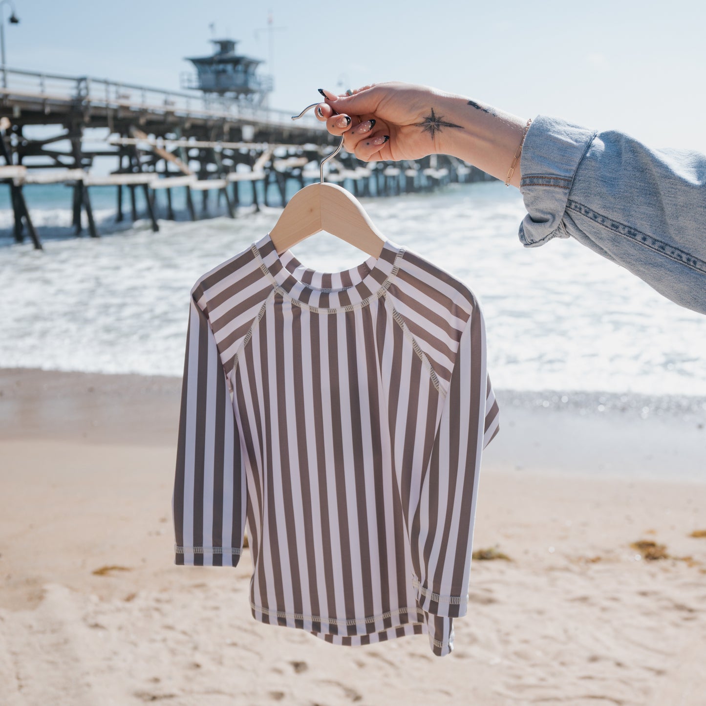 A hand holds the forever french baby Child Rash Guard | Olive Stripe—a long-sleeve, olive and white striped sun-protective top—by the beach with ocean and pier in the background under a clear blue sky.