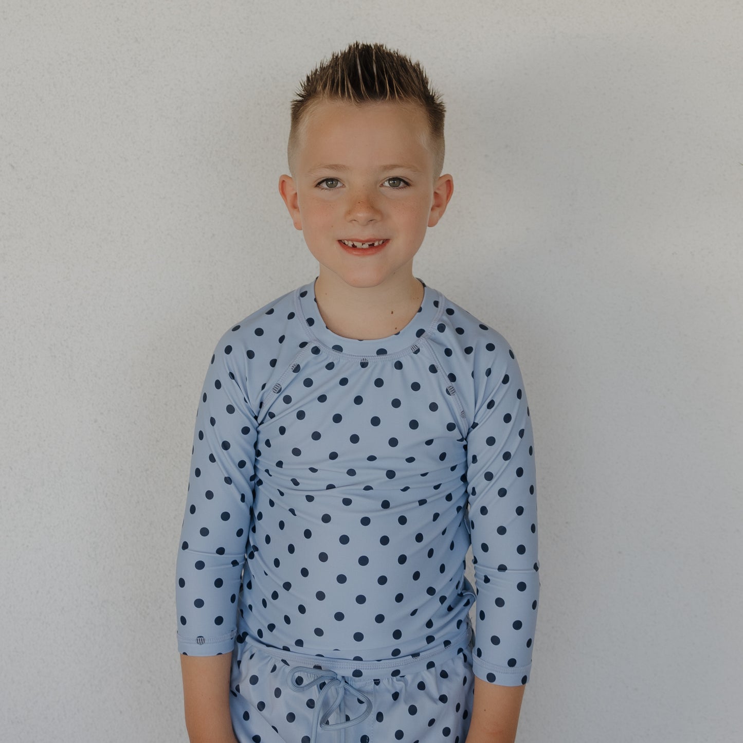 A young boy with short spiked hair smiles at the camera, wearing the forever french baby Child Rash Guard | Sully—light blue swimwear with dark blue polka dots and matching shorts—offering stylish sun protection against a plain light-colored wall.