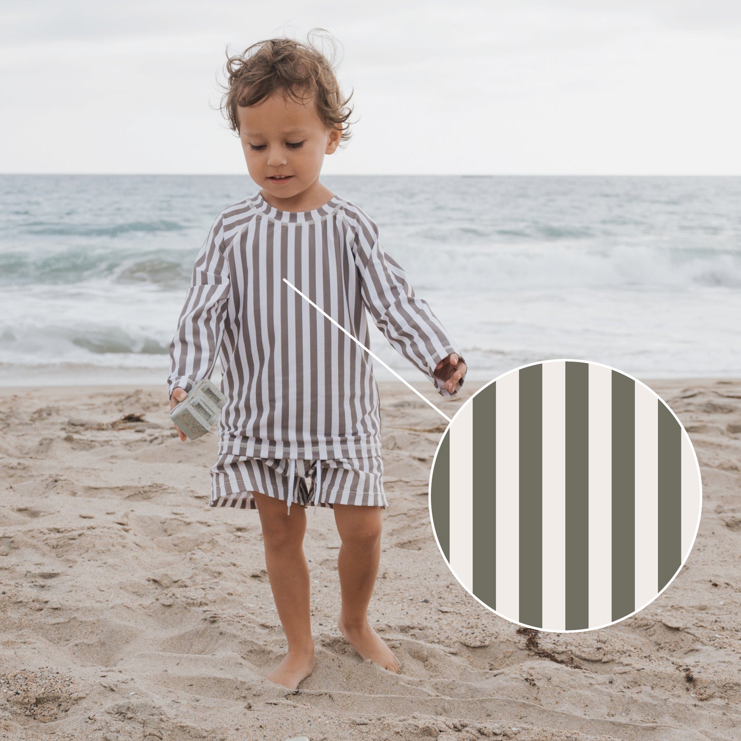 A young child wears the forever french baby Olive Stripe Rash Guard on the beach, featuring beige, gray, and white vertical stripes. A close-up shows the stripe details with waves in the background for a sun-protected look.