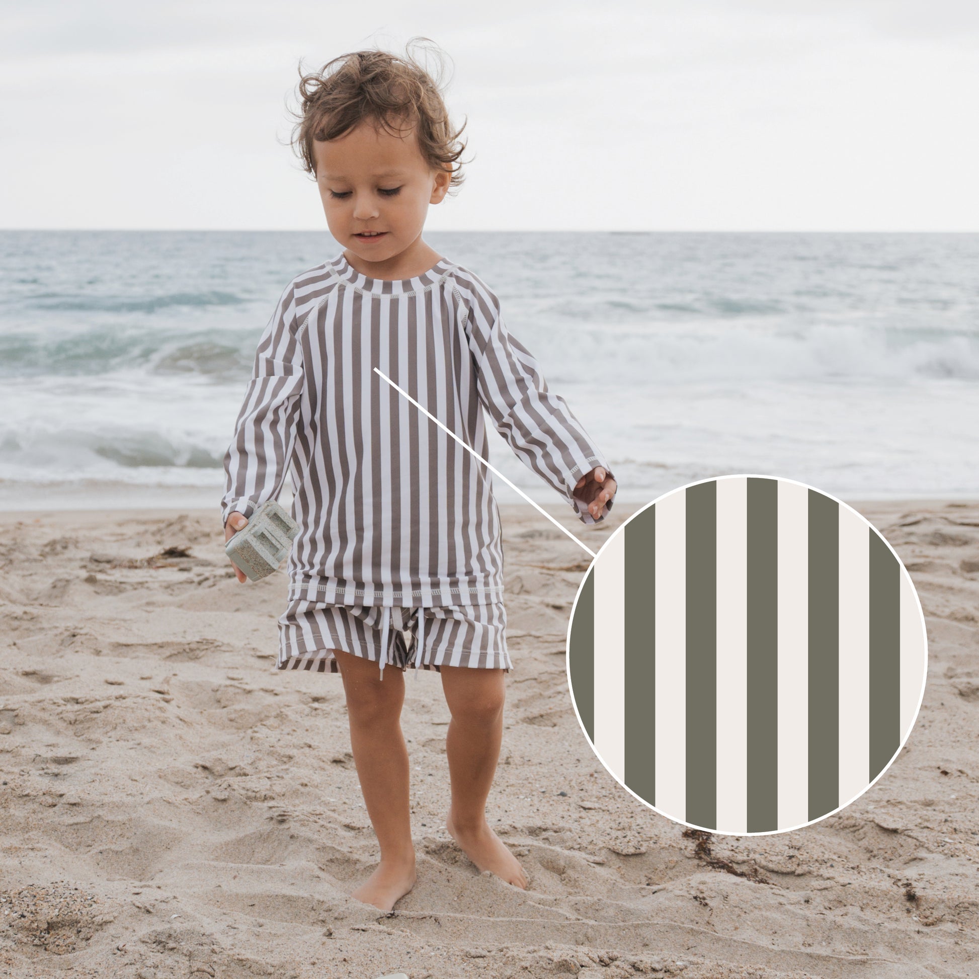 A young child wears the forever french baby Olive Stripe Rash Guard on the beach, featuring beige, gray, and white vertical stripes. A close-up shows the stripe details with waves in the background for a sun-protected look.