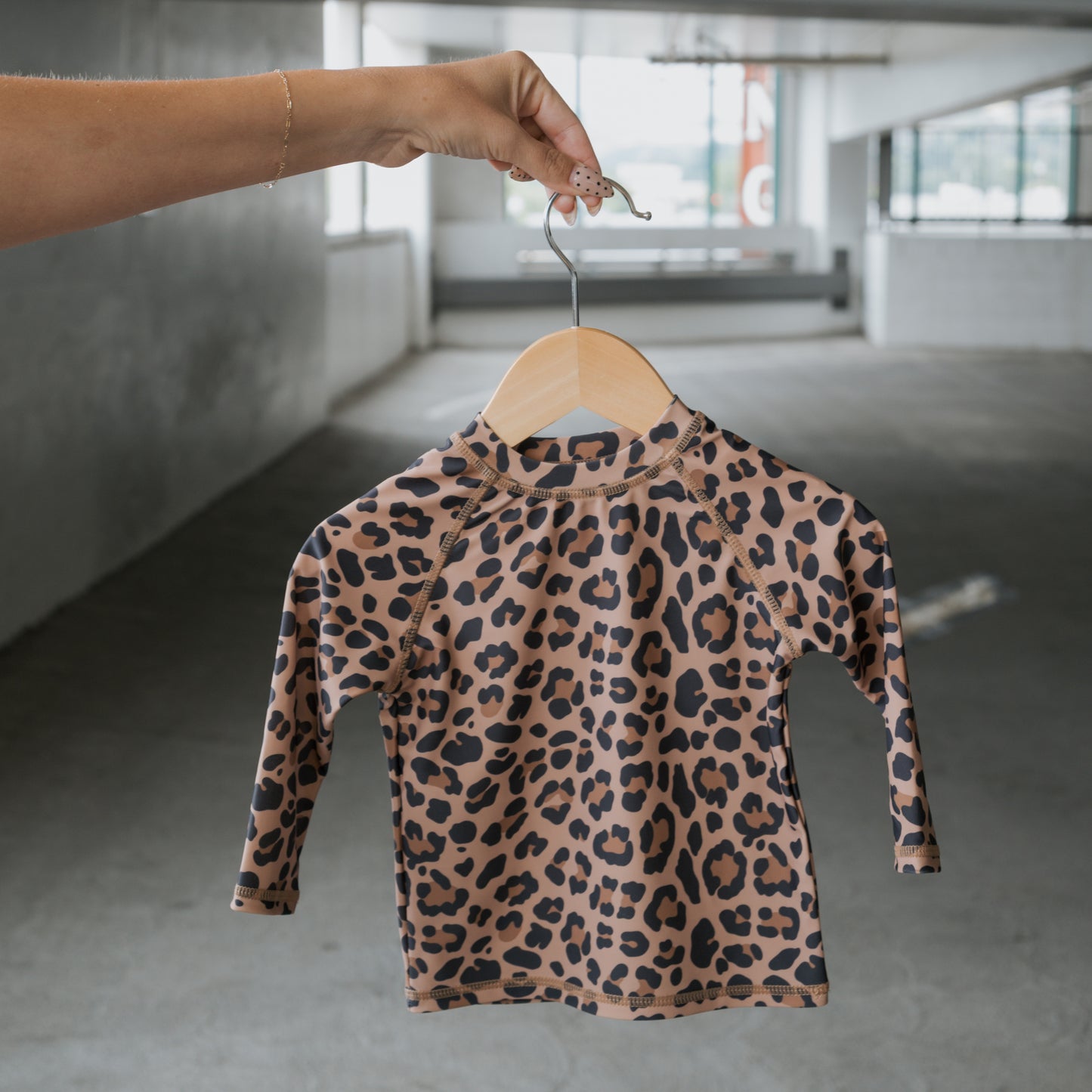 A hand holds up the *PRE-ORDER* Child Rash Guard | Leopard from forever french baby—a long-sleeved, leopard-print kids’ swim top—against a bright, blurred indoor background.
