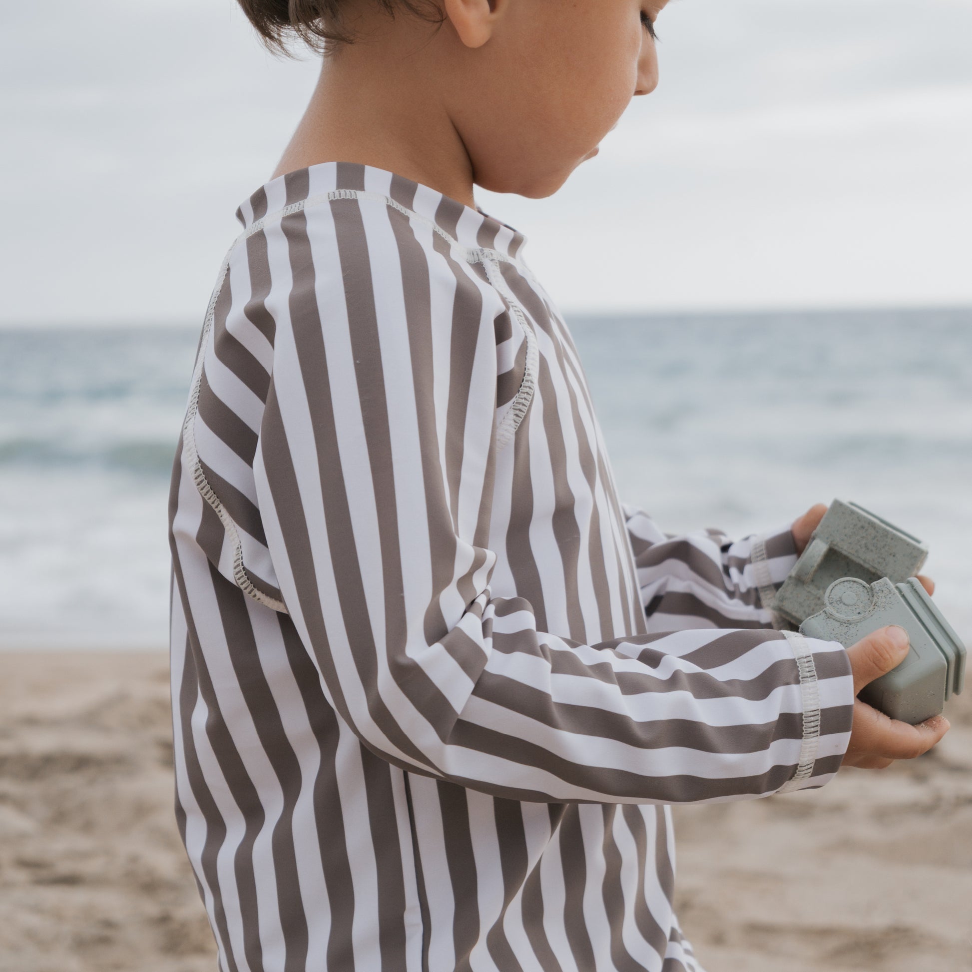 A young child wearing the forever french baby Child Rash Guard in Olive Stripe stands on a sandy beach holding a small gray toy camera, with the ocean and sky behind them, ready for fun and sun protection.