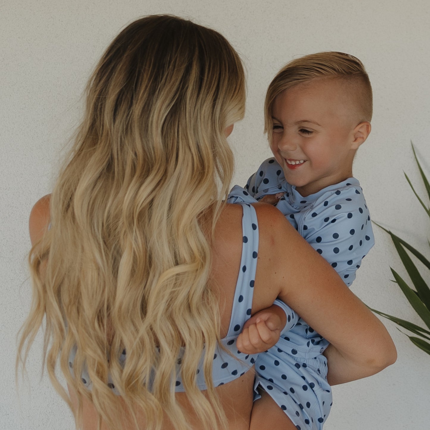 A woman with long, wavy blonde hair holds a smiling child wearing forever french baby’s Child Board Short | Sully. Both sport light blue family styles with dark blue polka dots, standing by a white wall and a green plant.