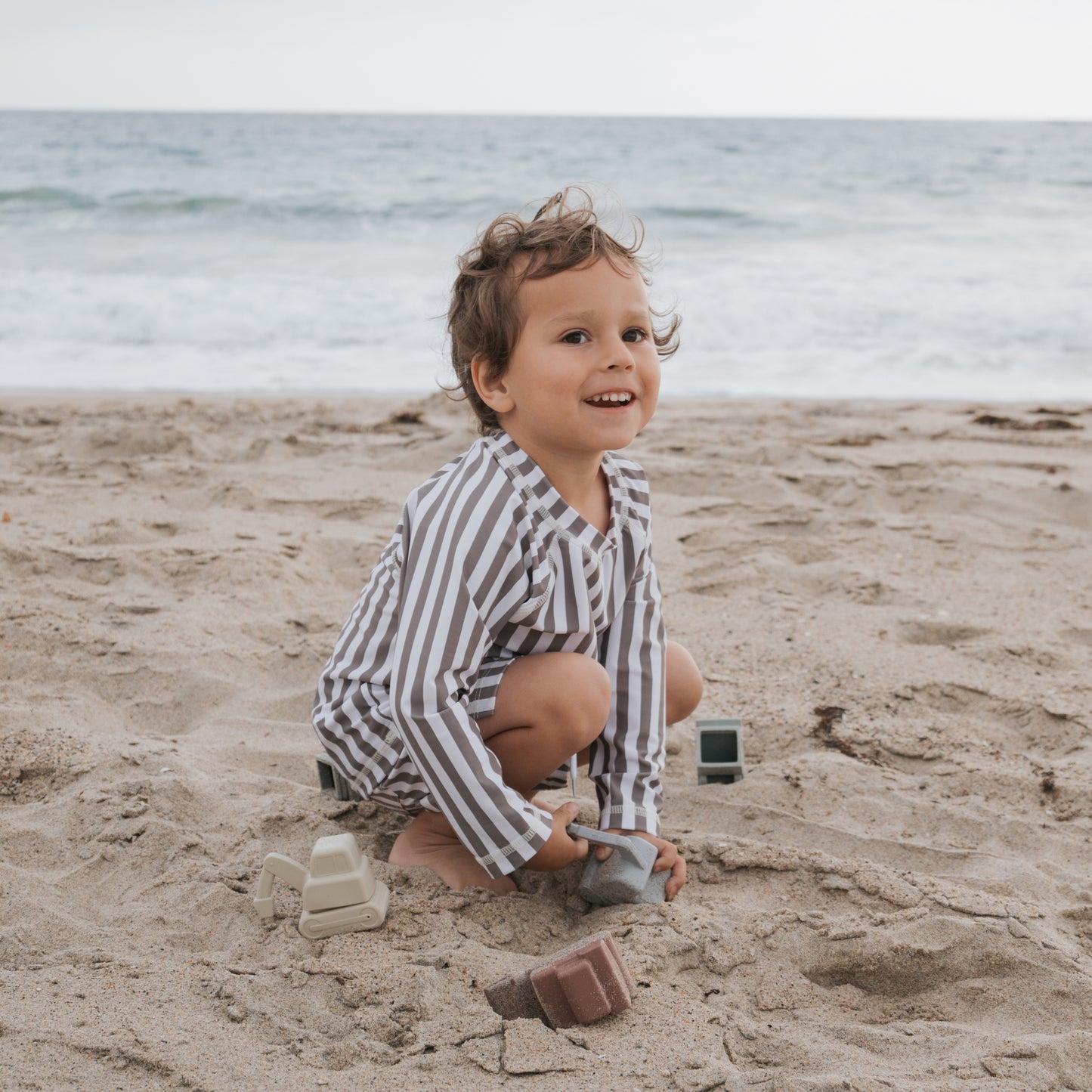A young child wearing the forever french baby Child Rash Guard | Olive Stripe smiles and plays with toy blocks on a sandy beach, with ocean waves behind them. The rash guard offers sun protection for carefree play under a cloudy sky.