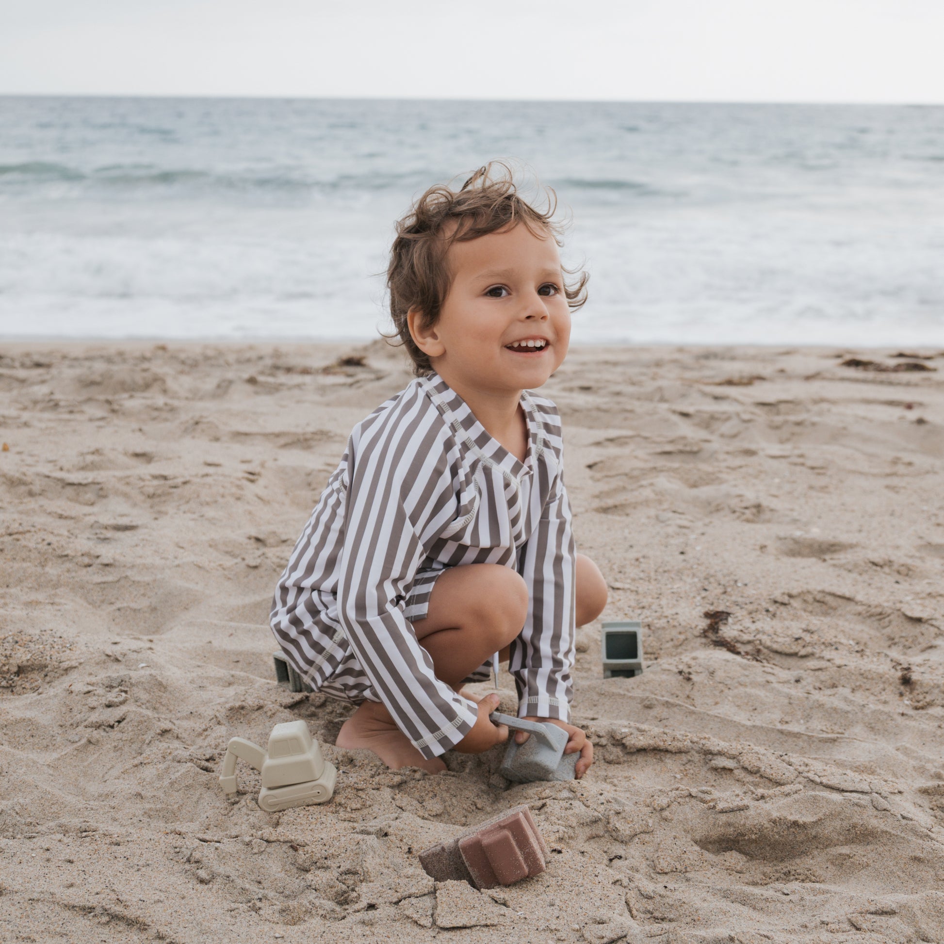 A young child wearing the forever french baby Child Rash Guard | Olive Stripe smiles and plays with toy blocks on a sandy beach, with ocean waves behind them. The rash guard offers sun protection for carefree play under a cloudy sky.