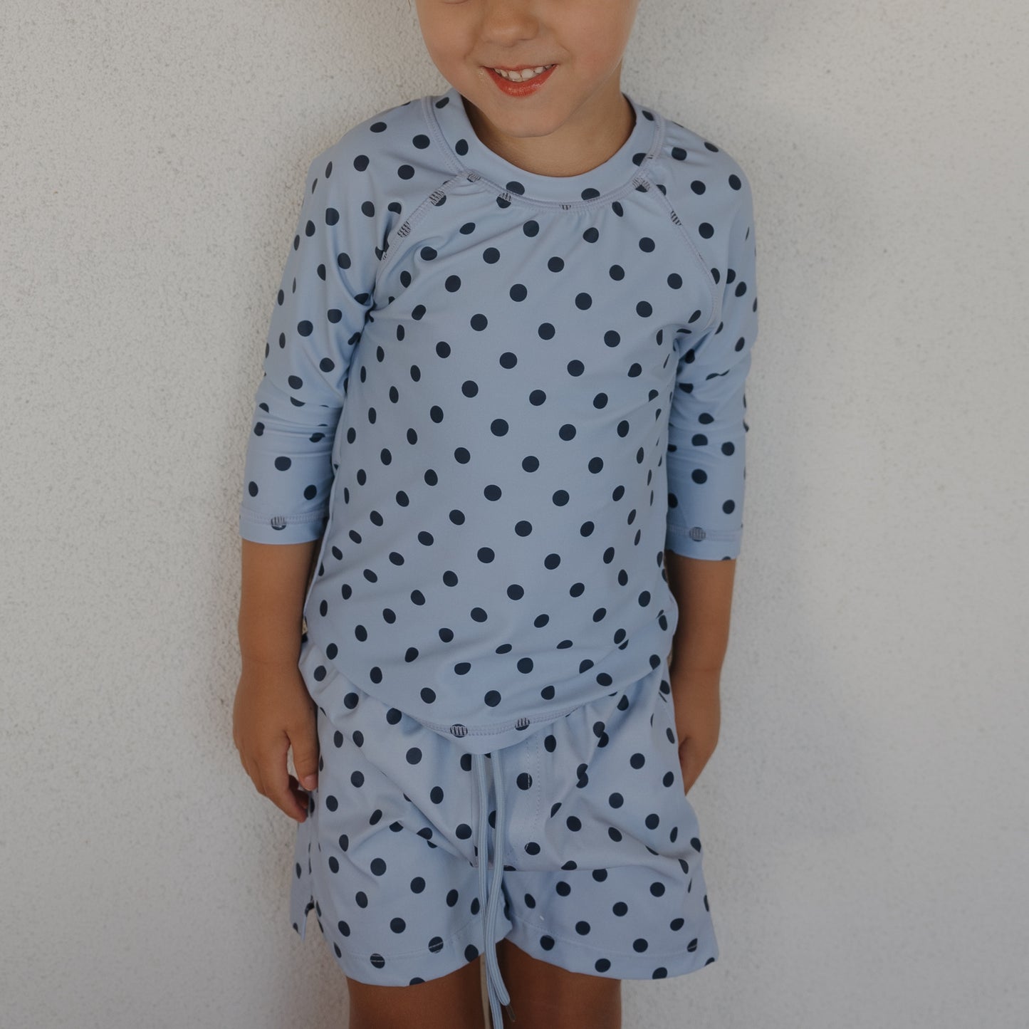A young child beams in the forever french baby Child Board Short | Sully set, a light blue long-sleeve top with navy polka dots and matching board shorts, standing against a plain white wall.
