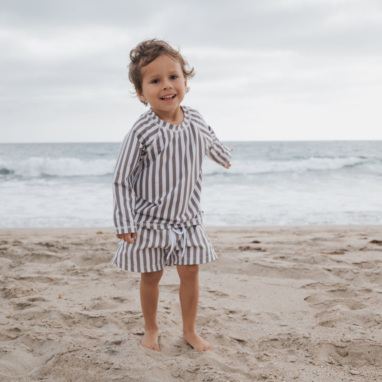 A young child wearing the forever french baby Child Rash Guard | Olive Stripe stands barefoot on the sandy beach, smiling with waves and clouds behind them, enjoying sun protection and comfort.