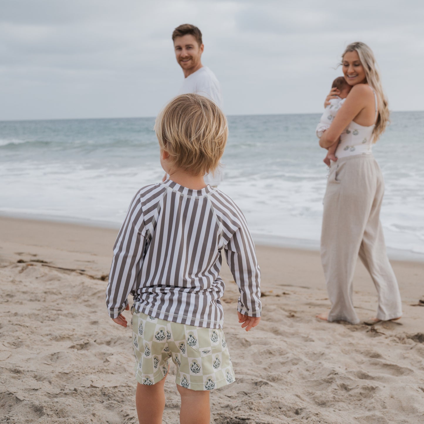 A young child in the forever french baby Child Rash Guard | Olive Stripe stands on the beach facing the ocean, as a smiling man and woman holding a baby look back at them. The sky is cloudy and waves roll toward the shore.