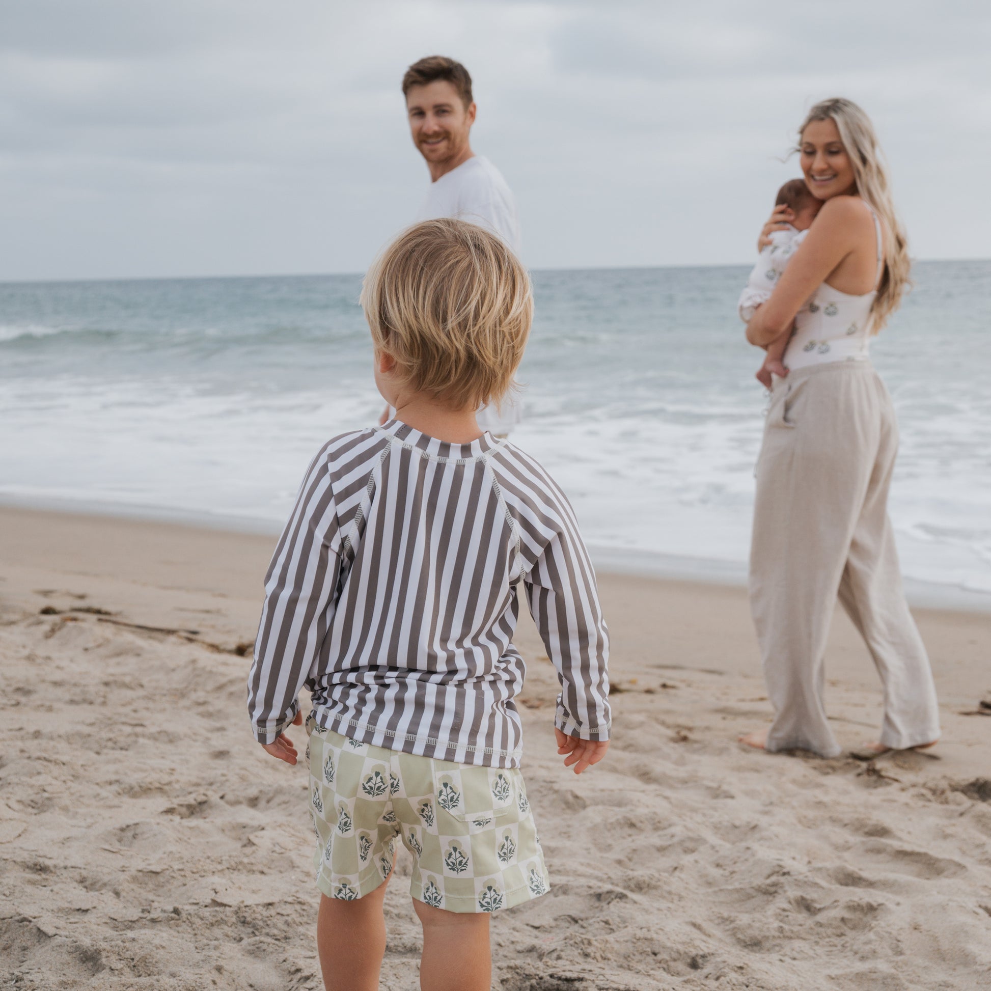 A young child in the forever french baby Child Rash Guard | Olive Stripe stands on the beach facing the ocean, as a smiling man and woman holding a baby look back at them. The sky is cloudy and waves roll toward the shore.