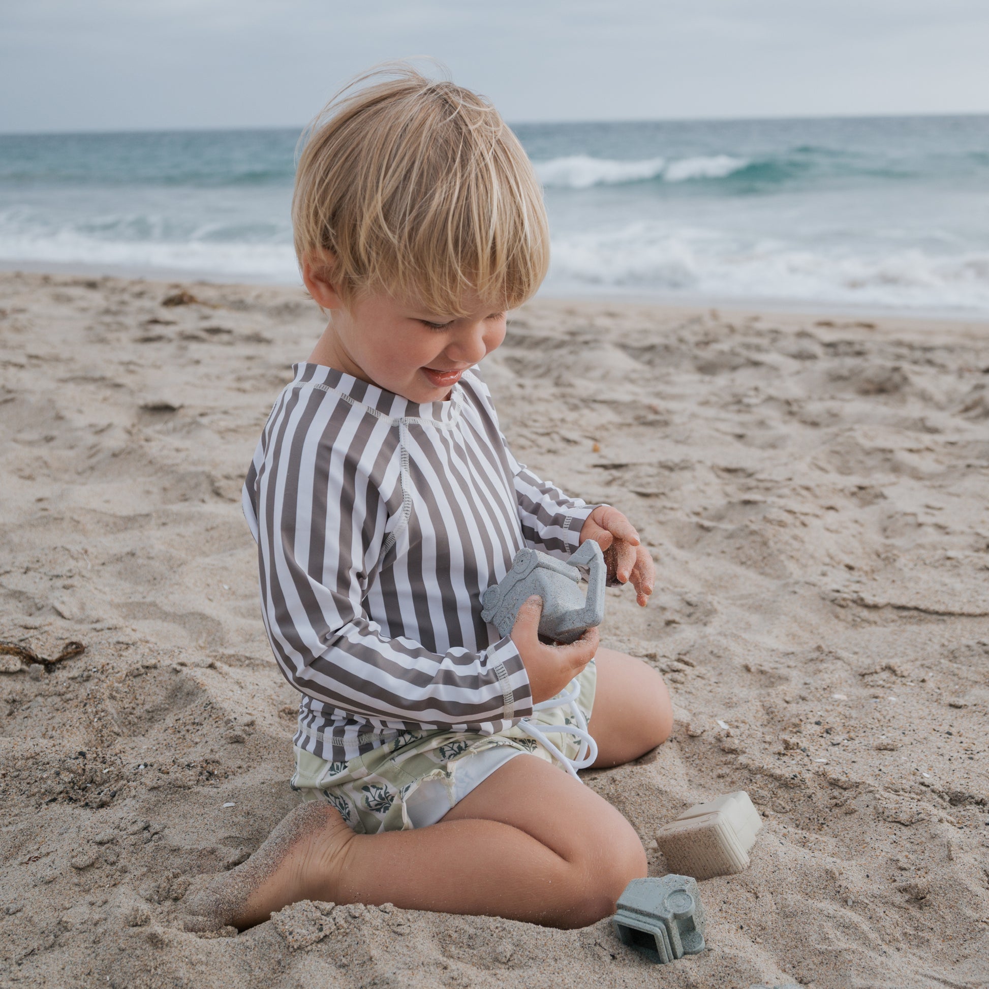 A young child wears the forever french baby Child Rash Guard | Olive Stripe while playing with toy buildings on a sandy beach, enjoying sun protection as part of their kids swimwear on a cloudy day.