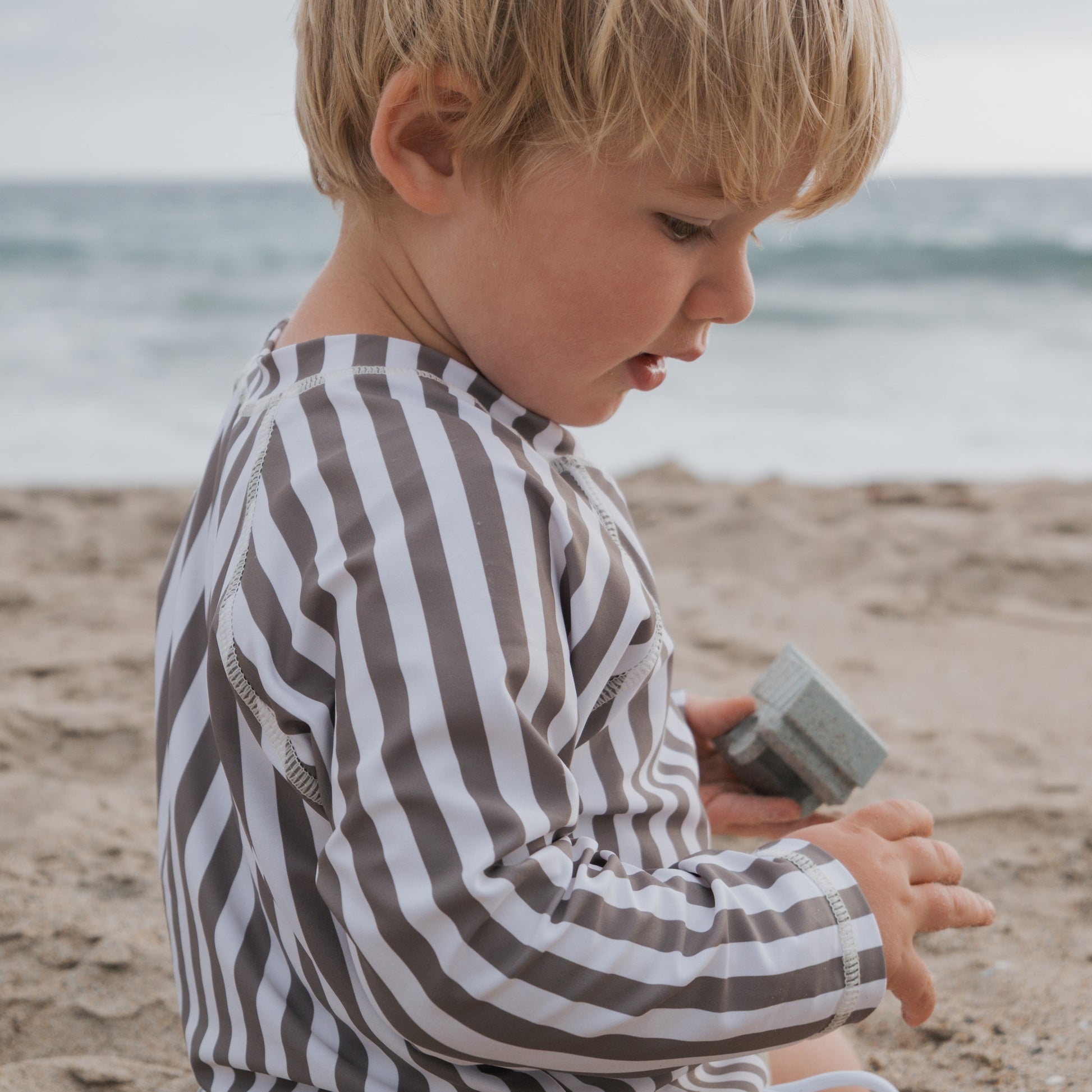A young child wears the forever french baby Olive Stripe Child Rash Guard while sitting on the sandy beach near the ocean, holding small toys and looking down at their hand.