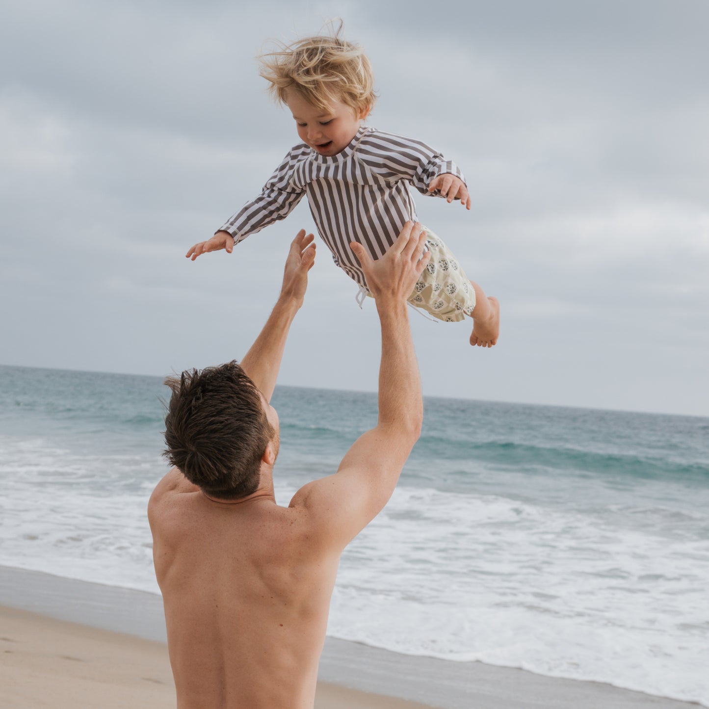 A man with short brown hair tosses a smiling child wearing the forever french baby Olive Stripe Child Rash Guard into the air on a sandy beach, waves and cloudy skies in the background.