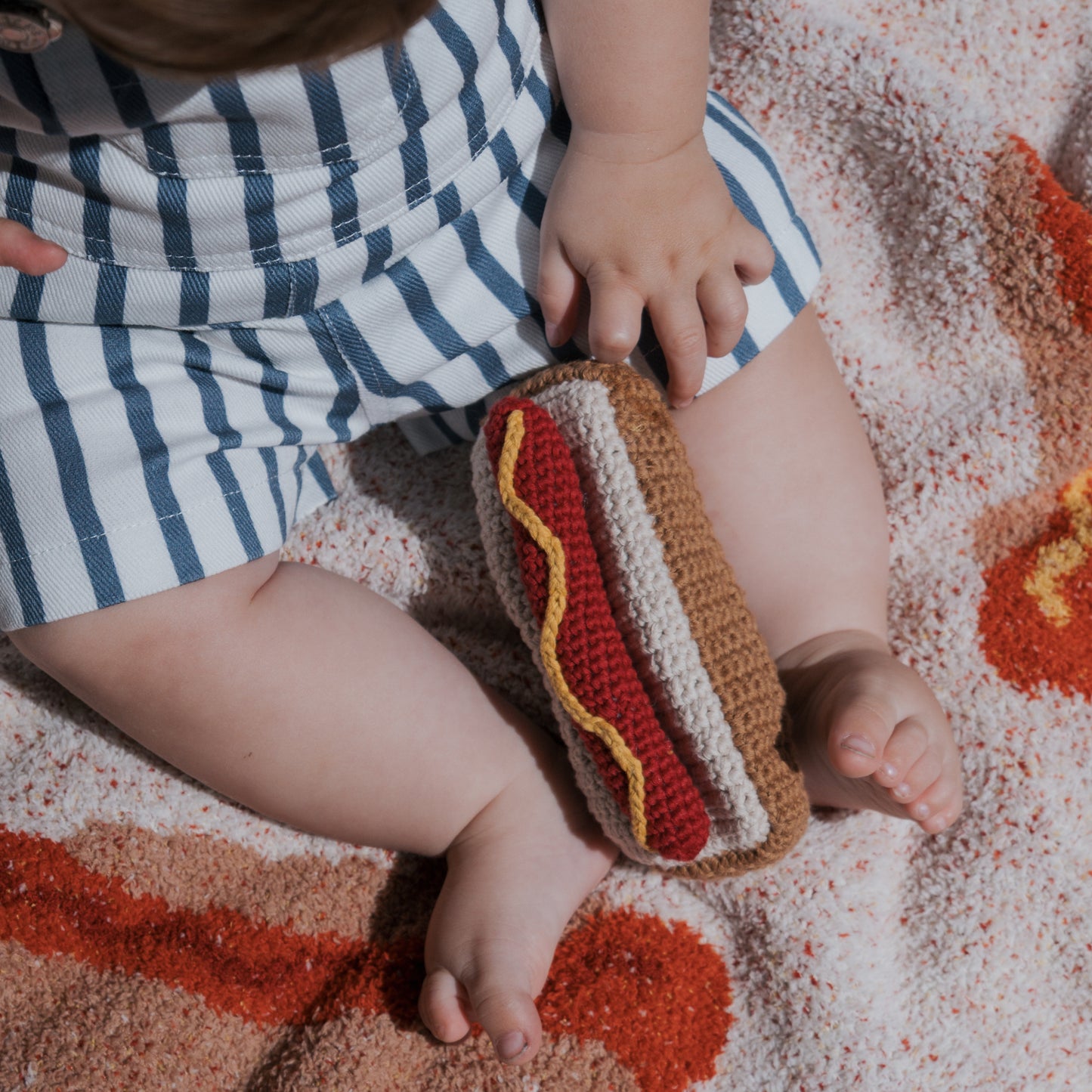 A baby in blue and white striped shorts sits on a forever french baby Plush Blanket | Hot Dogs, holding a knitted toy hot dog with mustard and ketchup details.