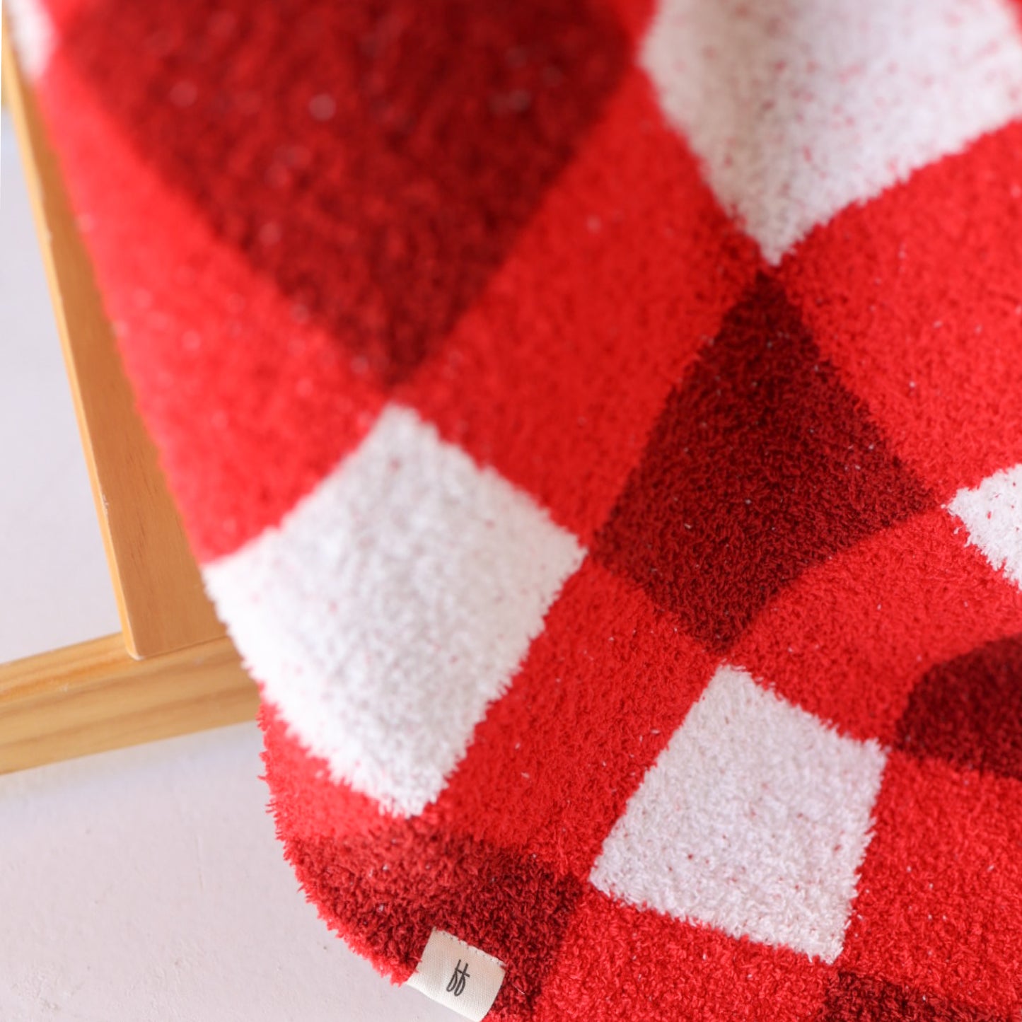 A close-up of the forever french baby Plush Blanket | Scarlet Gingham, featuring a red and white checkered texture and a small white tag near the edge, draped over a wooden surface.