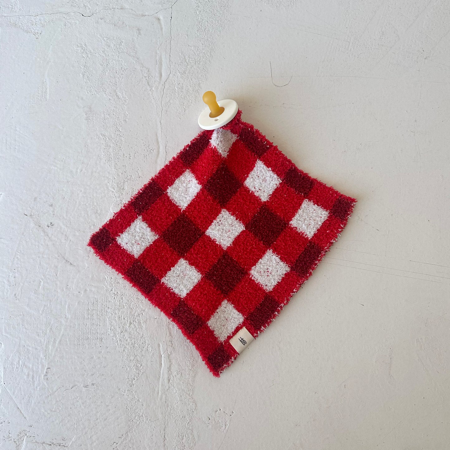 The Lovey | Scarlet Gingham by forever french baby, a red and white checkered microfiber blanket with a pacifier attached at one corner, is displayed flat on a textured white surface.