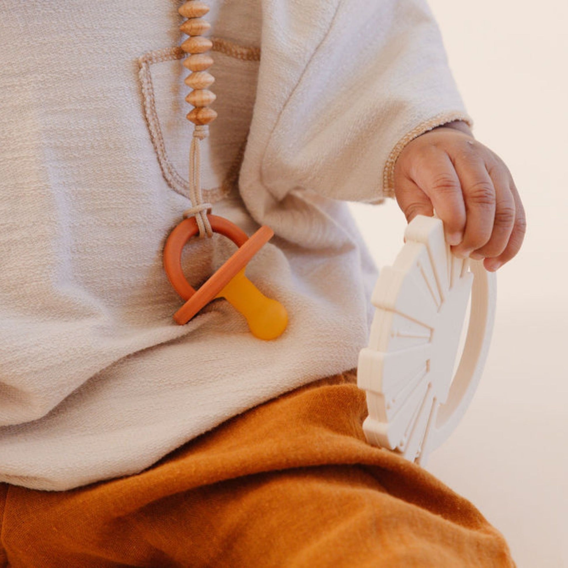 A child wearing a light-colored top and mustard pants holds a white circular toy in one hand. The child has the *Modern Pacifier | Caramel* by forever french baby, attached to a string and clipped to their shirt. The image focuses on the child's midsection and hands, highlighting only part of their body along with key baby accessories.