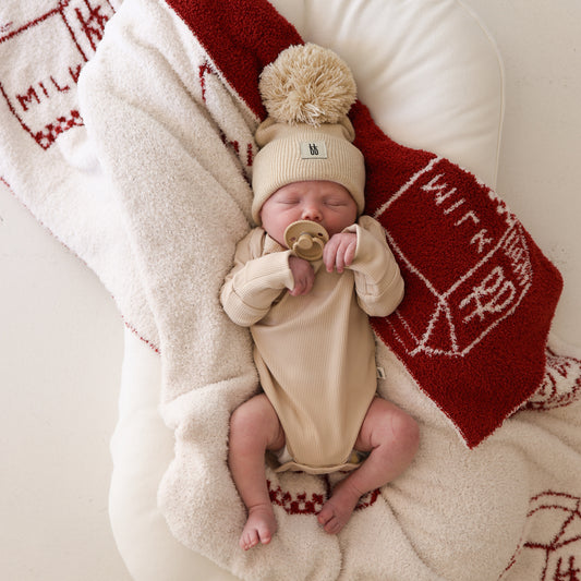 A newborn in the forever french baby ff Pom Top Knit Beanie in Oatmeal lies on a soft cream blanket, holding a pacifier. Nearby is a red and cream blanket with milk carton designs, made from breathable yarn.