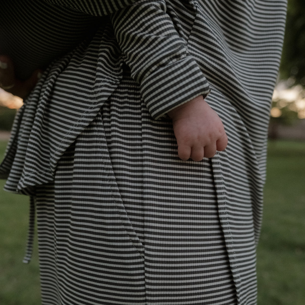 A close-up of a hand resting at the side, wearing the forever french baby Women’s Bamboo Button Up Lounge Set in Forest Stripe Ribbed, with a soft grassy background out of focus.