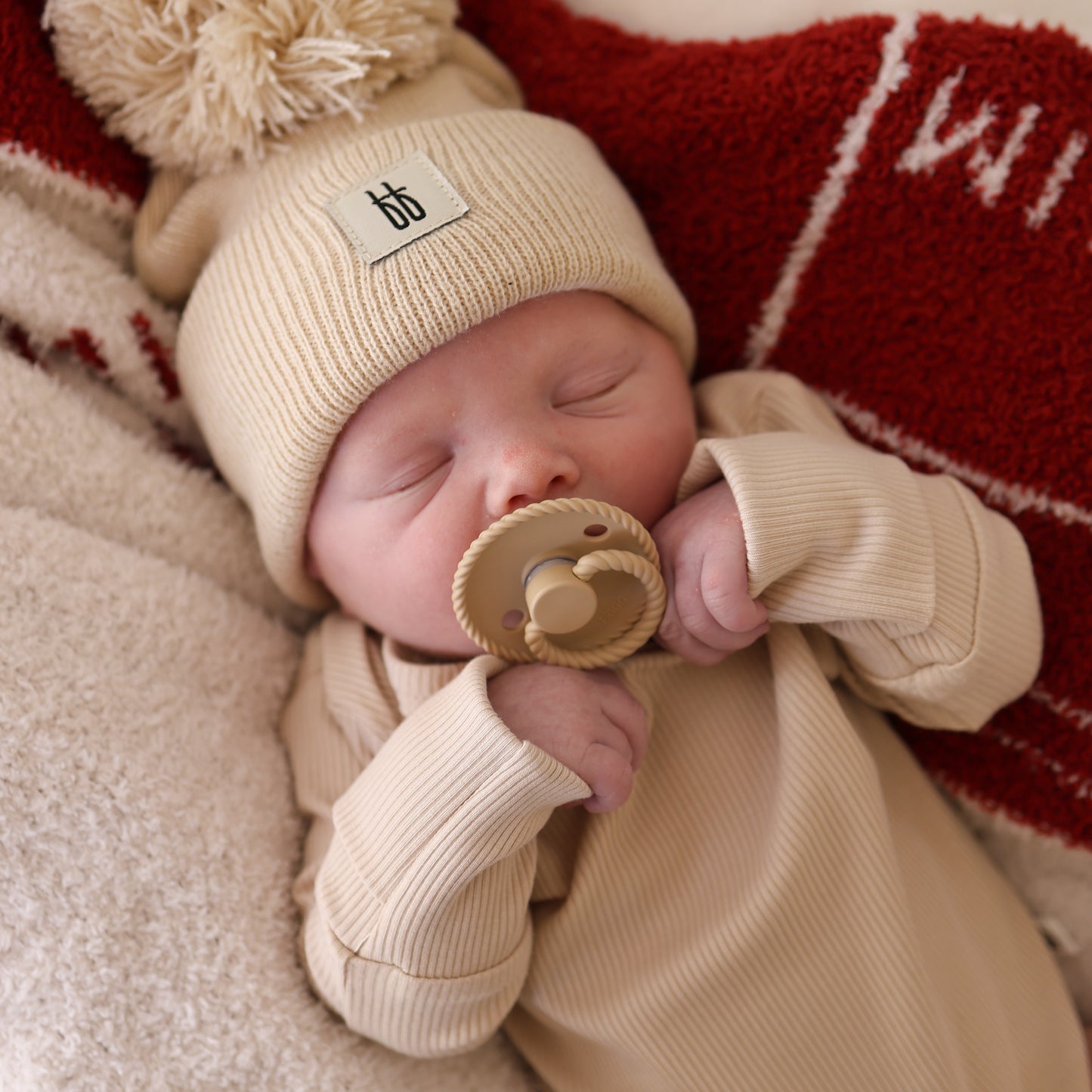 A baby sleeps on a red and white blanket, holding a tan pacifier, dressed in the forever french baby Long Sleeve Bamboo Snapsuit | Ribbed Cream and a knit pom-pom hat—hypoallergenic for comfort on delicate skin.