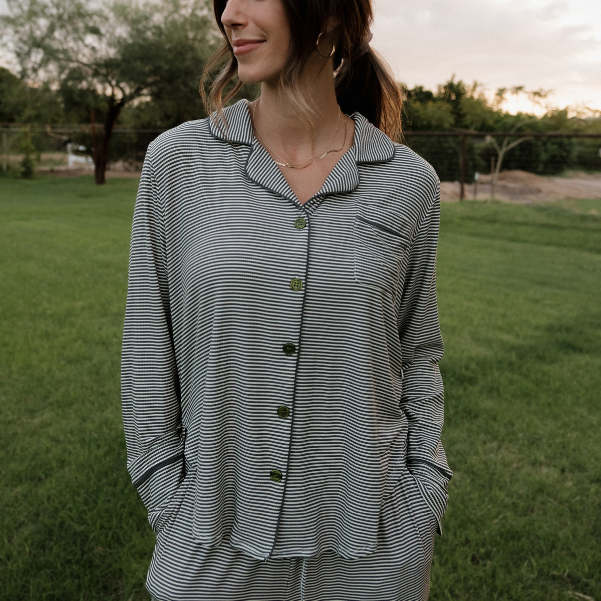 A woman stands outside on the grass, wearing the forever french baby Women’s Bamboo Button Up Lounge Set in Forest Stripe Ribbed, with her hands in her pockets. Trees and a fence appear in the background. Her face is partially turned away and she has a slight smile.