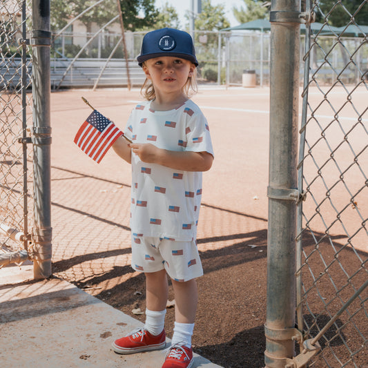 A young child stands by a chain-link tennis court fence, wearing the forever french baby Bamboo Short Set | Americana Ribbed, paired with a navy hat, red sneakers, and holding a U.S. flag.