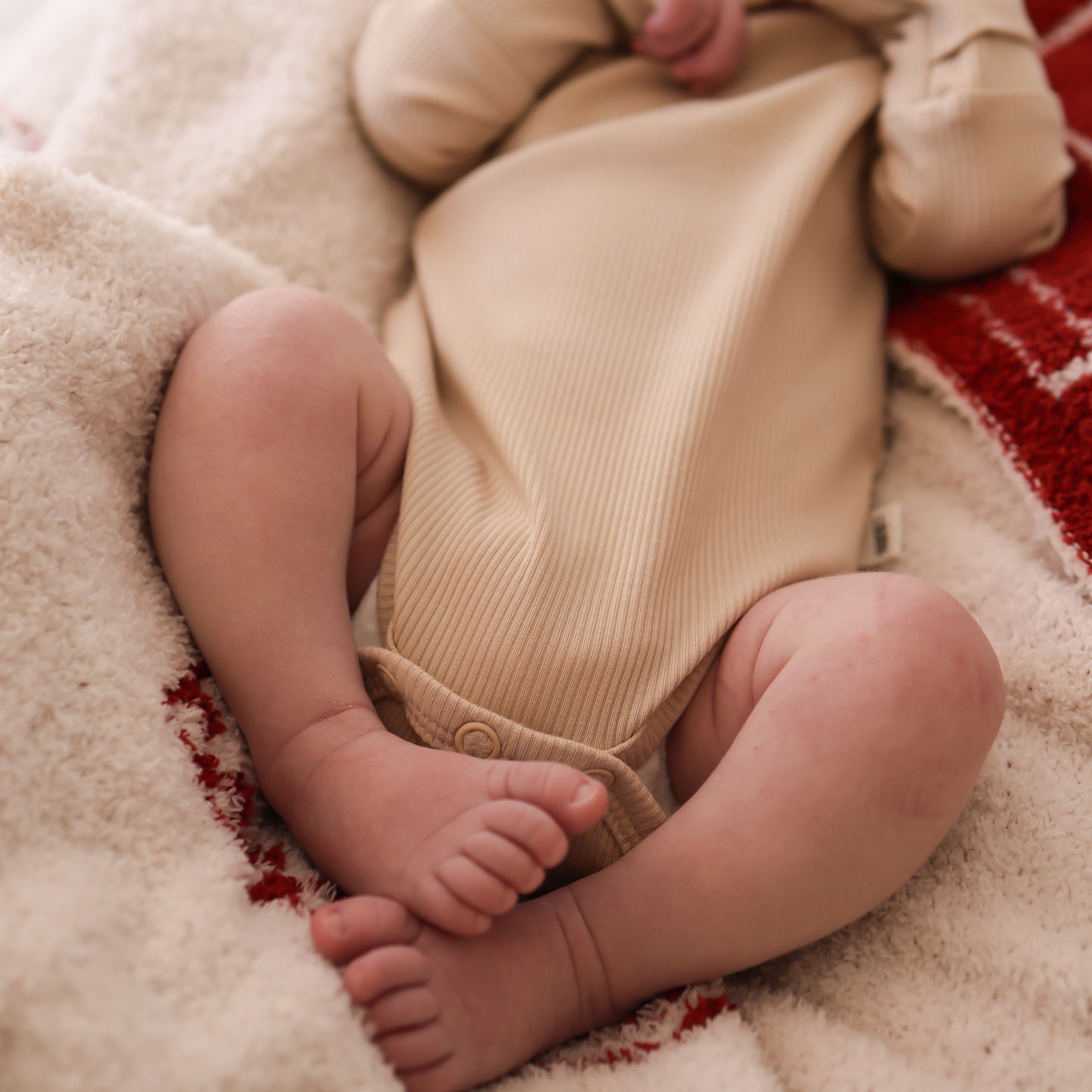 A baby wears the forever french baby Long Sleeve Bamboo Snapsuit in Ribbed Cream, lying on a cozy white and red blanket with their little legs and feet showing.