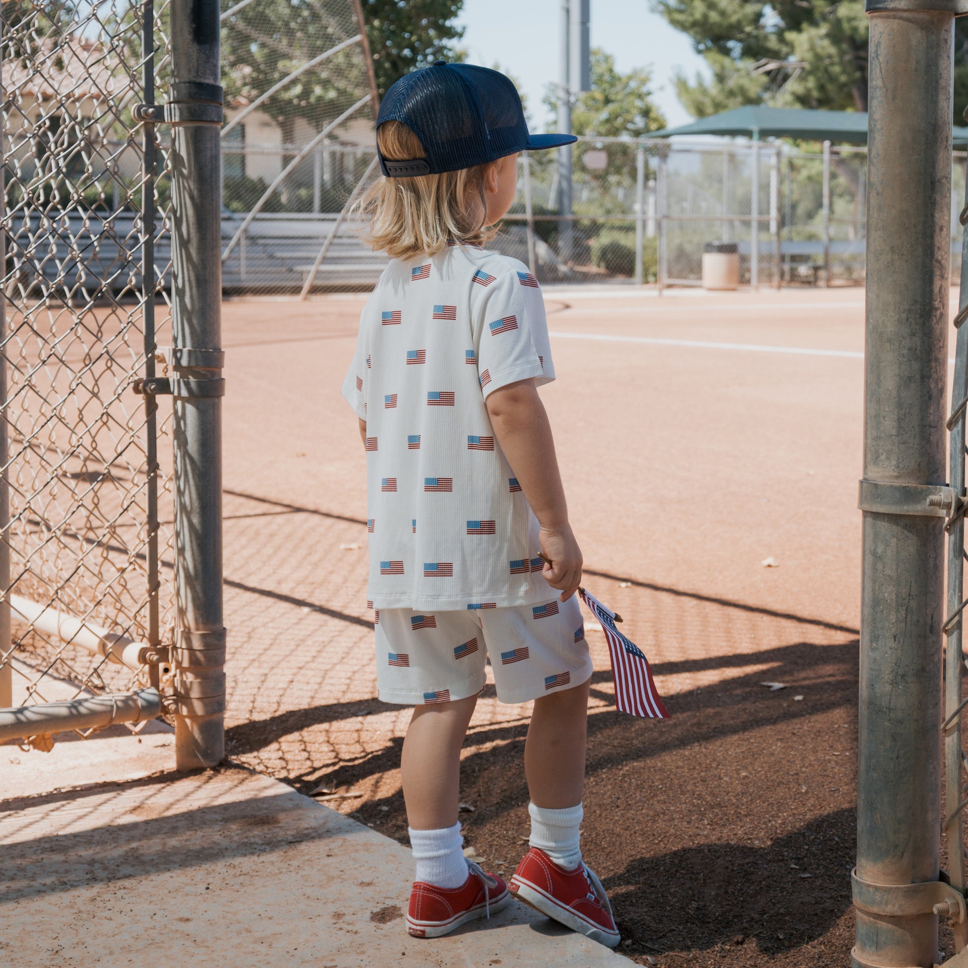 A young child in the forever french baby Bamboo Short Set | Americana Ribbed stands by a baseball field fence, holding a small American flag and wearing red sneakers and a blue cap.