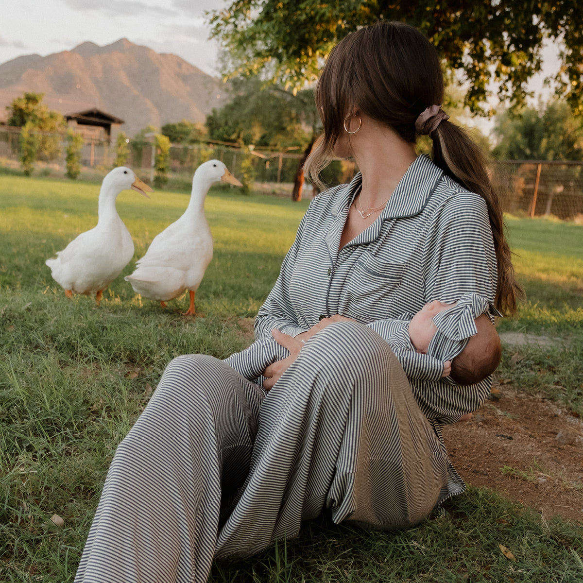 A woman wearing the forever french baby Women’s Bamboo Button Up Lounge Set in Forest Stripe Ribbed sits on the grass holding a baby, watching two white ducks strolling nearby in a lush outdoor setting with mountains in the background.