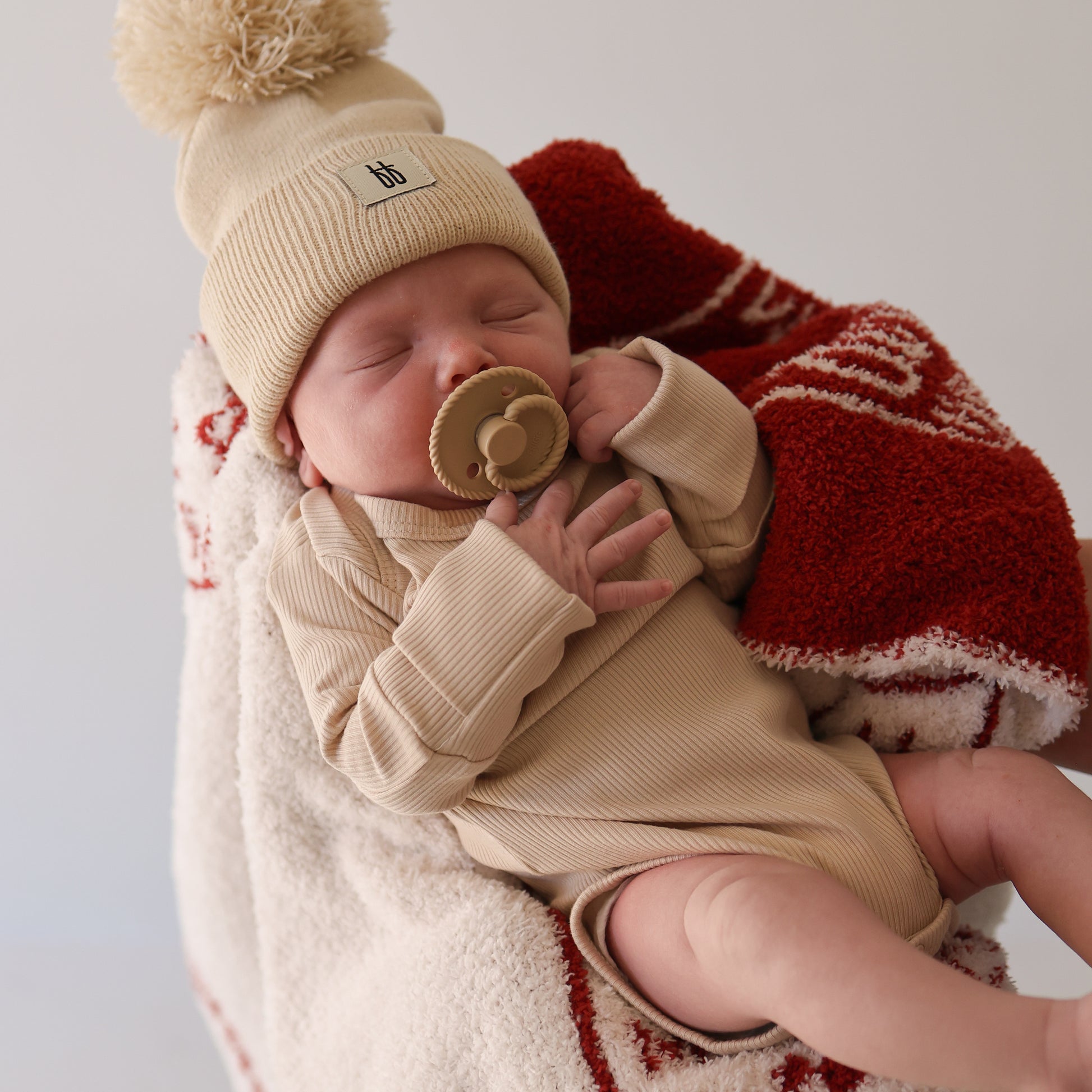 A newborn asleep with a pacifier, wrapped in a red and white blanket, wears forever french baby’s Long Sleeve Bamboo Snapsuit | Ribbed Cream and a beige pom-pom beanie for hypoallergenic comfort.