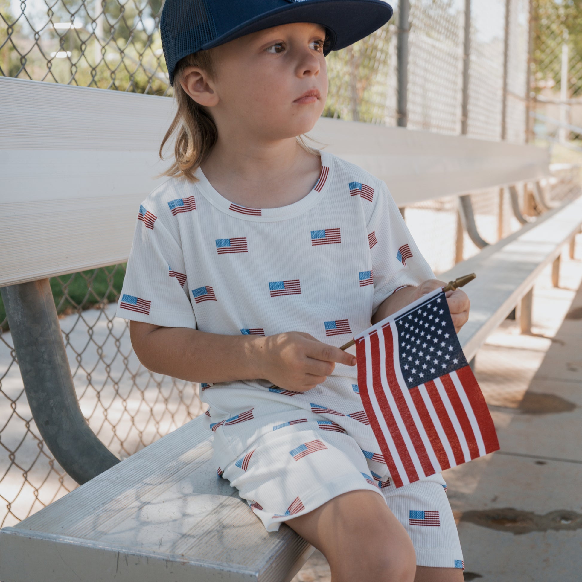 A young child in the forever french baby Bamboo Short Set | Americana Ribbed—a blue, American flag print outfit and cap—sits on a bench by a chain-link fence, holding a small flag on a sunny day.