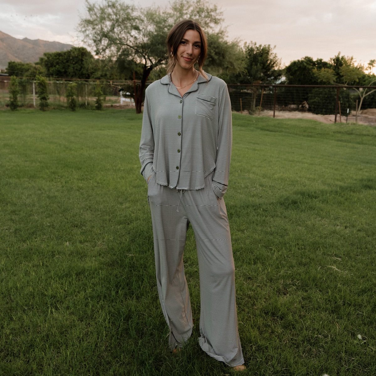 A woman wearing the forever french baby Women’s Bamboo Button Up Lounge Set in Forest Stripe Ribbed stands on a grassy lawn with trees and mountains in the background, her hands in her pockets and a slight smile on her face.