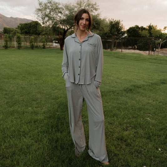 A woman wearing the forever french baby Women’s Bamboo Button Up Lounge Set in Forest Stripe Ribbed stands on a grassy lawn with trees and mountains in the background, her hands in her pockets and a slight smile on her face.